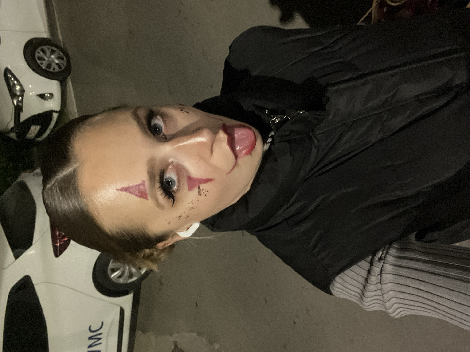 A person is posing for a selfie at night in a paved area. The person is wearing a black puffy jacket and a grey and black striped shirt. They have pale skin, light eyes, and dark hair pulled back. Red makeup is applied to their face: a triangle on their forehead, dots under their eyes, and what appears to be a smear of blood on their tongue. A partial view of two white cars is visible in the background, including the letters "MC" on the side of one. There are no definitive elements to confirm the location is Boreti, Montenegro.