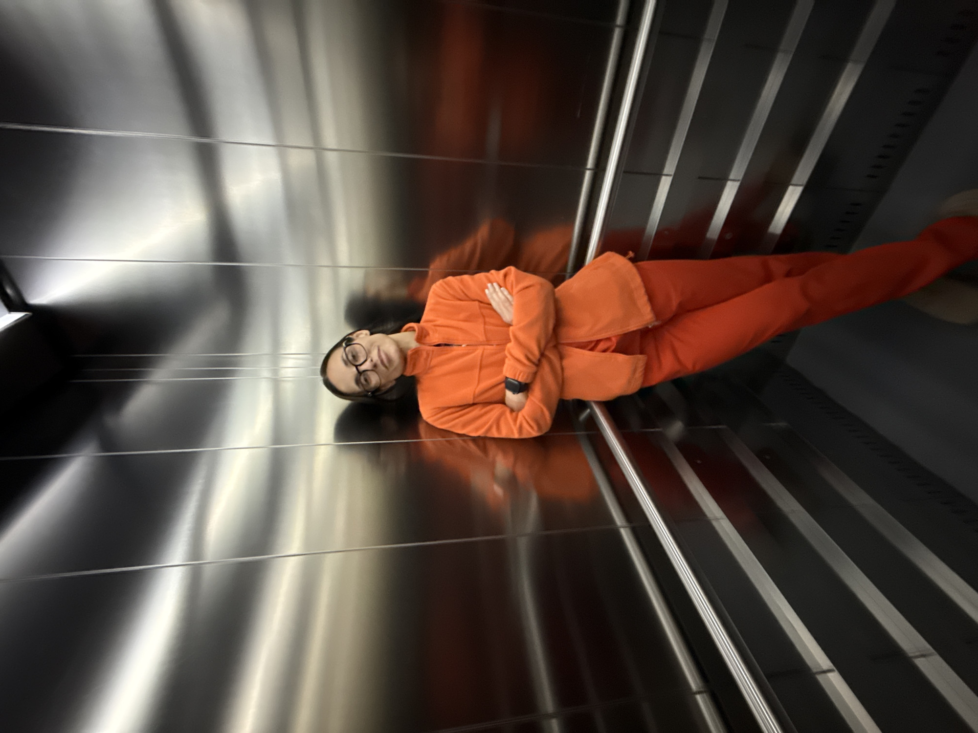 A young person is captured in a well-lit, modern interior space, likely an elevator or a similarly enclosed area. They are wearing a vibrant orange two-piece outfit consisting of a fleece jacket with a zipper and matching pants. Their arms are crossed, and a black smartwatch is visible on their left wrist. They have short, dark hair and are wearing round-rimmed glasses, looking directly at the camera with a slight, relaxed smile.
The setting features highly reflective, metallic silver walls, one of which shows a blurred, elongated reflection of the person. Another wall, behind the subject, is characterized by vertical stripes of varying silver and dark gray tones, with subtle, unreadable markings or indicators along its length. The floor is also reflective, mirroring the person's form. The dominant colors in the image are the striking orange of the attire and the cool, reflective silver and gray tones of the environment. The lighting is internal and uniform, highlighting the metallic surfaces. No discernible text is visible within the frame. The image appears to be taken in Pleven, Bulgaria.
