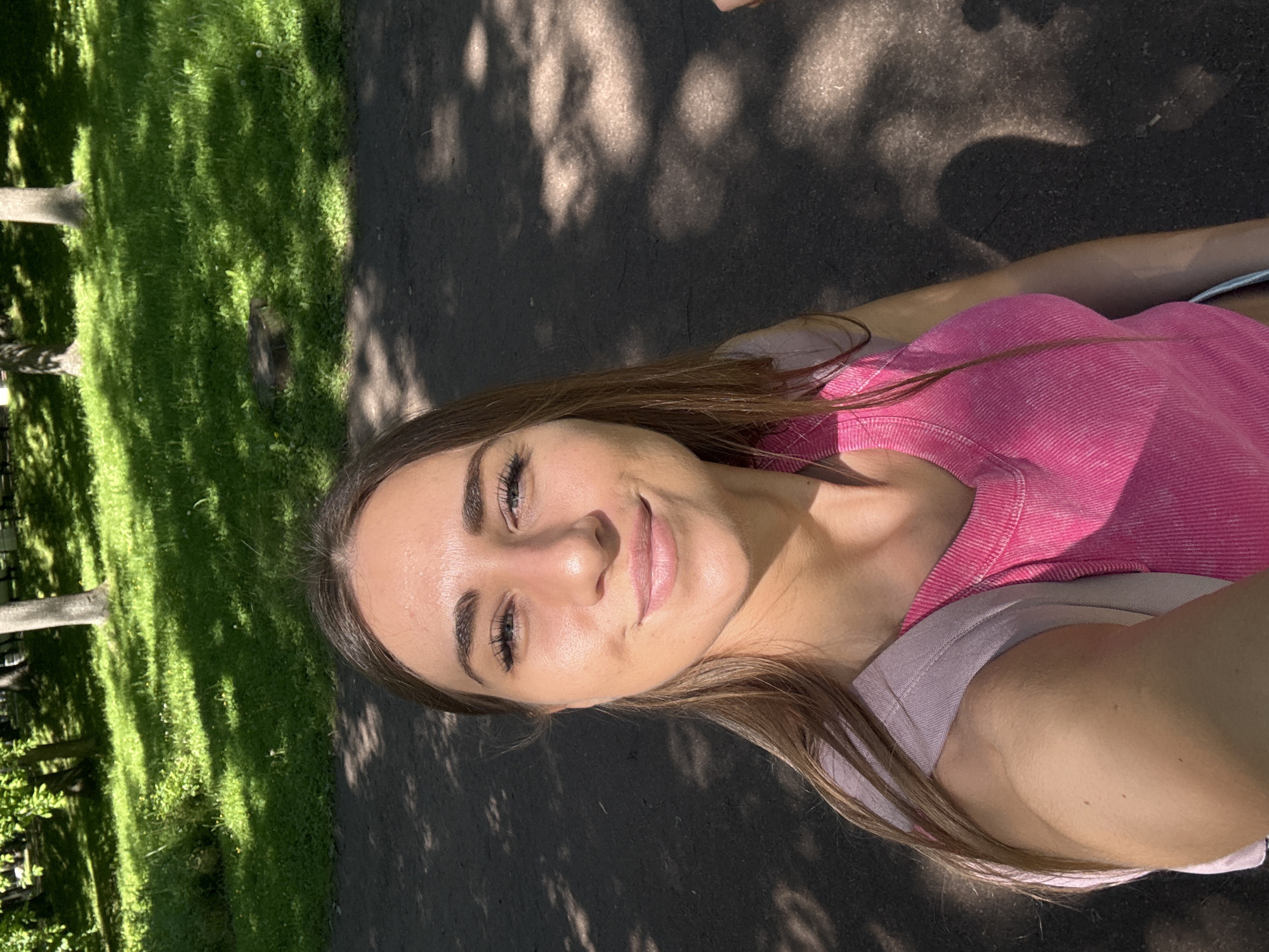 The image captures a female individual taking a selfie in a park in Saint Petersburg, Russia, under bright daylight conditions. The woman, positioned in the lower two-thirds of the frame, has long, dark brown hair and fair skin with sunlit highlights on her forehead, nose, and cheeks. She has defined eyebrows and visible eyelashes, and is looking directly at the camera with a closed-mouth smile. She wears a ribbed, fuchsia-pink sleeveless top. A light lavender or pale pink strap, consistent with a backpack or shoulder bag, is visible over her right shoulder.
The environment behind her consists of a dark grey paved path that occupies the mid-ground and extends to the foreground on the left. This path is marked by sharp, elongated shadows from unseen overhead foliage, indicating a sunny day. Beyond the path, the background features a lush green lawn interspersed with lighter green patches and small white wildflowers, notably dandelions. Several mature trees with green leaves are visible in the distance, also casting shadows across the grass. A tree stump is discernible on the edge of the grassy area to the left of the frame. The overall lighting suggests a clear sky and direct sunlight.