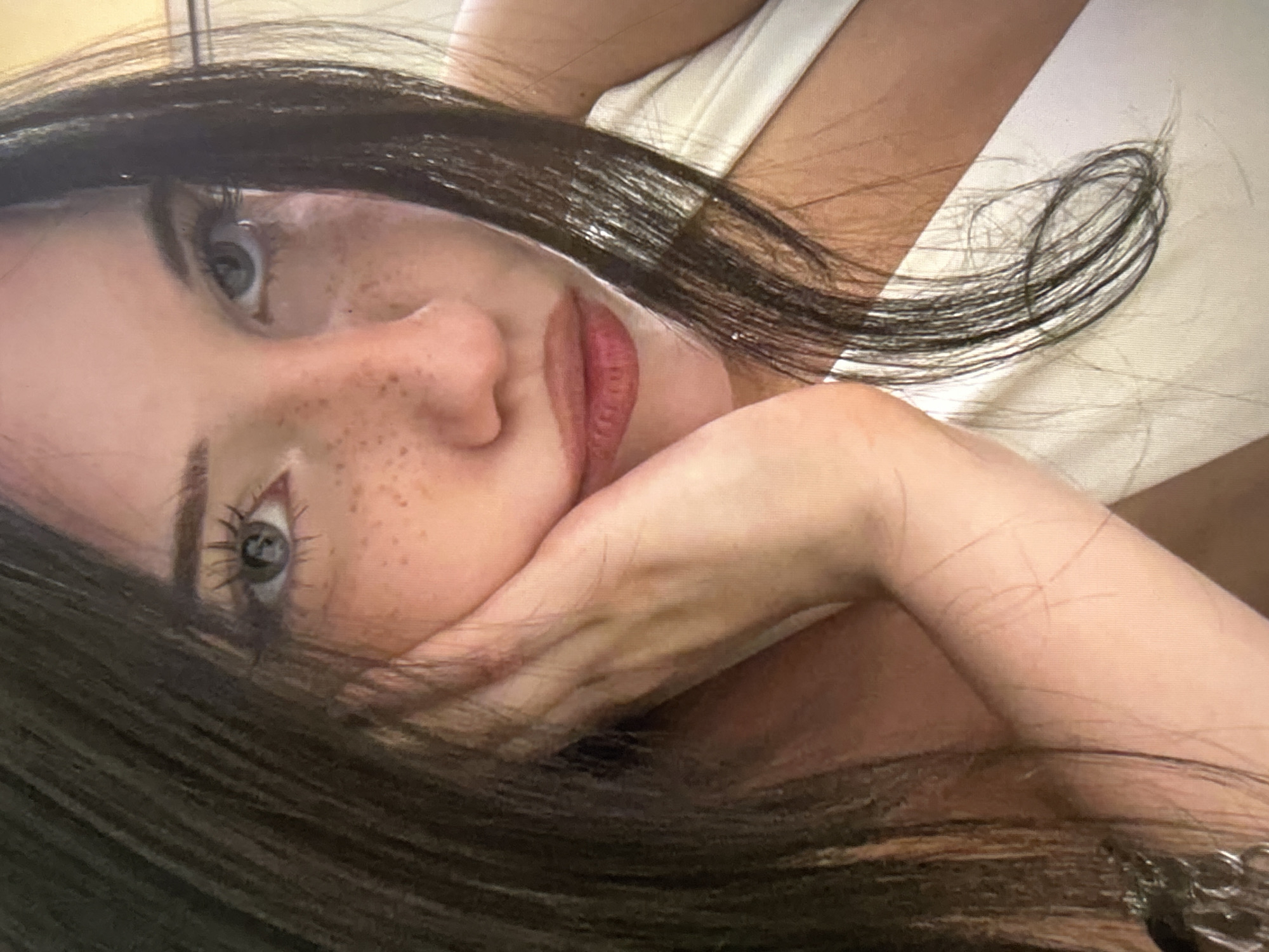 The image is a close-up, high-angle, portrait-style shot of a young woman with long, dark brown hair. She has fair skin with freckles across her nose and cheeks. Her eyes are light blue and she is looking upwards. Her left hand is resting on her cheek, with her fingers slightly curled. She is wearing a white top. The background is indistinct but appears to be indoors. The image does not provide any information about the location in the city of Cupra Marittima, Italy.