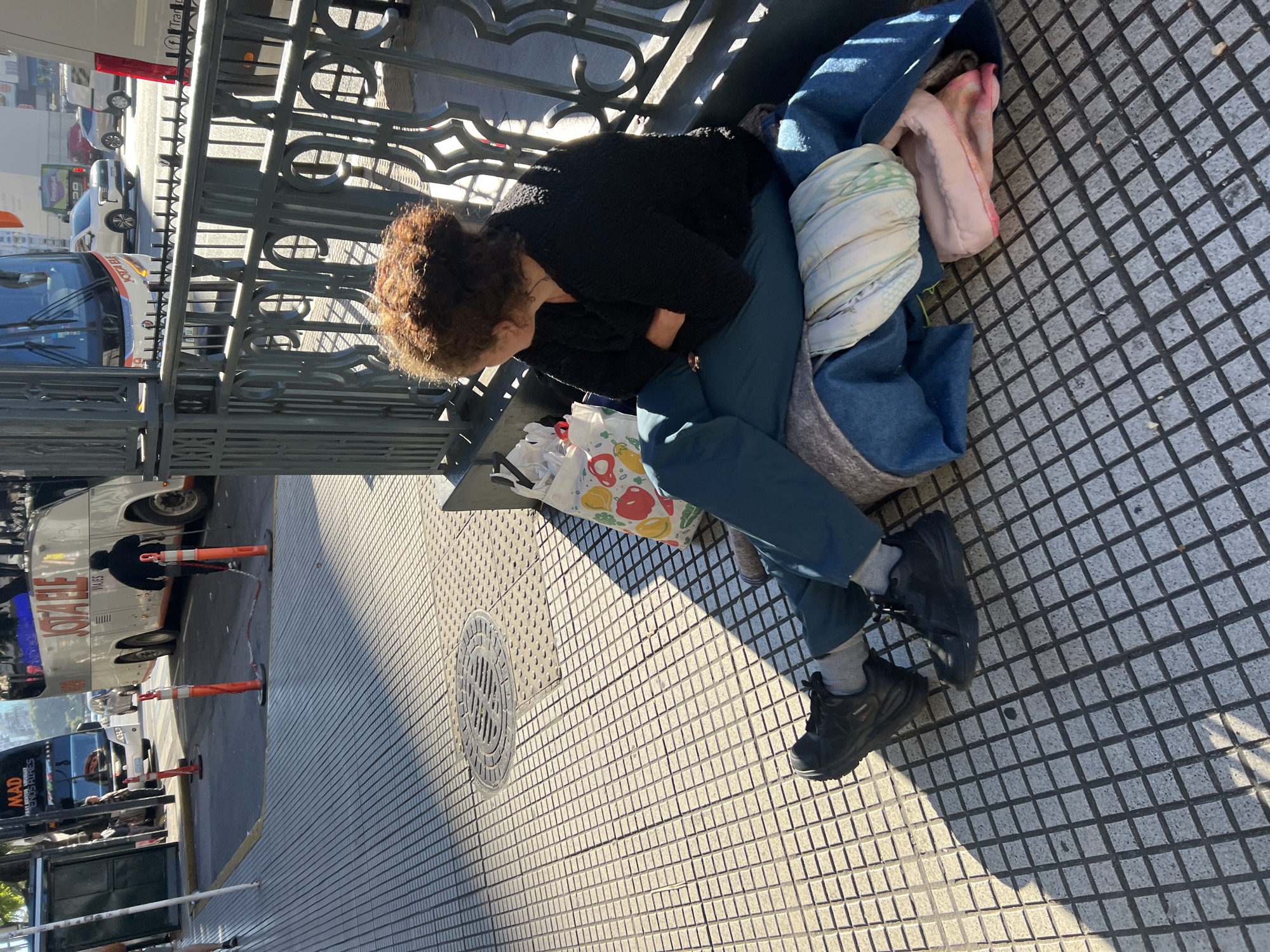 A woman with curly hair is sitting on a bench in Buenos Aires, Argentina. She is wearing a black sweater, blue pants, and black sneakers. She has her arms crossed and is leaning forward. Next to her is a bag and some blankets. The bench is located on a sidewalk with a patterned tile floor. A metal railing separates the bench from the street, where buses and cars can be seen. The photograph is taken in bright daylight.