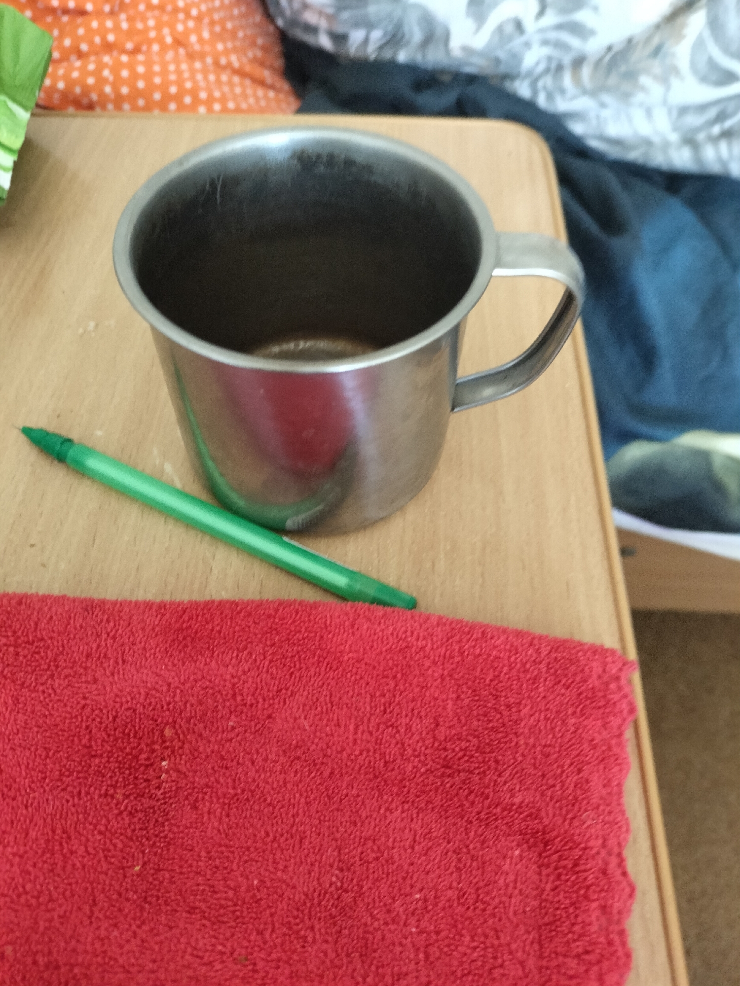 A close-up, eye-level shot shows a metallic mug with a handle, a green pen, and a red terrycloth towel on a light brown wooden surface. The mug is open at the top and appears empty. The pen is positioned diagonally across the surface, with its tip pointing towards the left edge of the frame. The red towel occupies the lower portion of the image, with its textured surface clearly visible. In the background, out of focus, are hints of bedding with orange and white polka dots, and a dark blue fabric. The scene appears to be indoors, possibly in a bedroom or living area. The image does not contain any specific landmarks or environmental cues that would allow for the identification of its location within the city of Chernihiv, Ukraine.