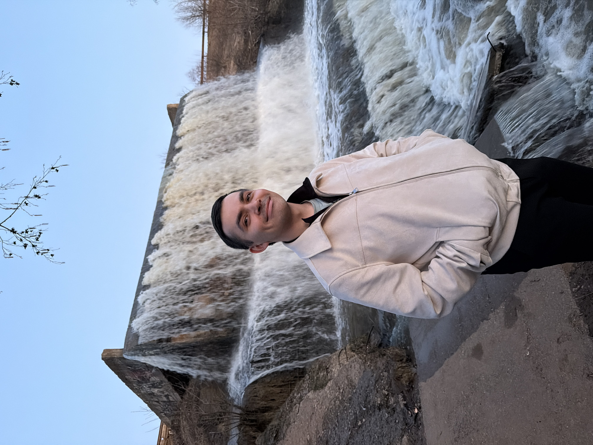 The image features a man standing outdoors in front of a multi-tiered waterfall or dam spillway. The man, positioned slightly to the right of the frame, is wearing a light-colored, possibly beige or off-white, zipped jacket with a collar, over a lighter-toned inner shirt and dark pants. He has dark hair, looks towards the camera, and displays a faint, pleasant smile. His hands appear to be clasped together or tucked into his jacket at waist level.
Behind the man, a significant volume of white, foamy water cascades down several concrete or stone steps, indicating a strong flow. The sky above is a clear, light blue, suggesting good weather conditions during the daytime. Bare tree branches are visible on the left side of the foreground and in the distance above the waterfall, hinting at a colder season such as late fall, winter, or early spring. The ground where the man stands appears to be a paved or concrete path. The overall setting is a scenic outdoor location, likely near a dam or a natural waterfall, in Kamennoye, Russia. There is no visible text in the image.