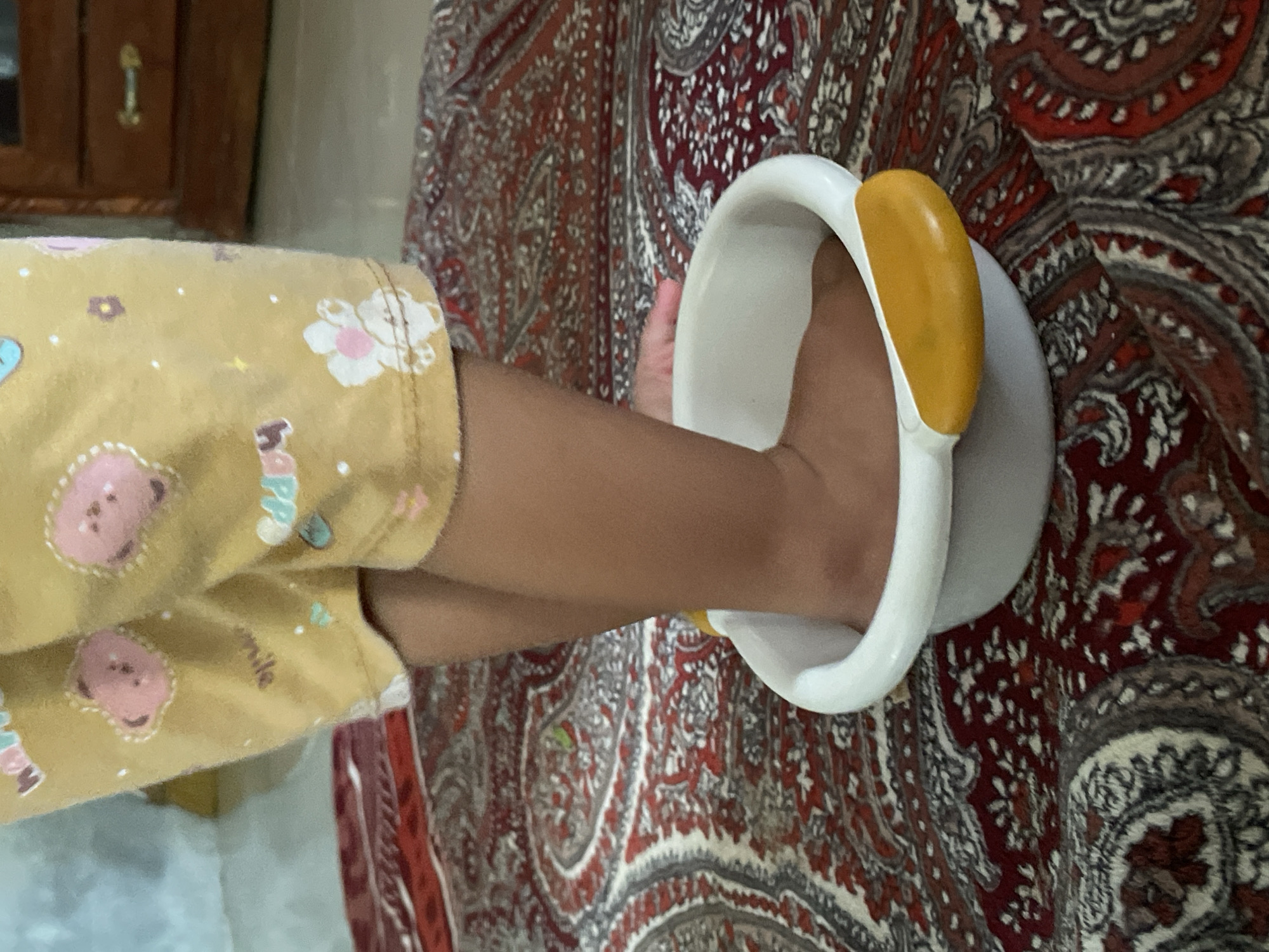 A child's bare foot is inserted into a white and yellow plastic bowl on a patterned reddish brown and gray textile surface. The child's arm and pajama-clad leg are also visible to the left of the bowl. The location is not discernible from the information.