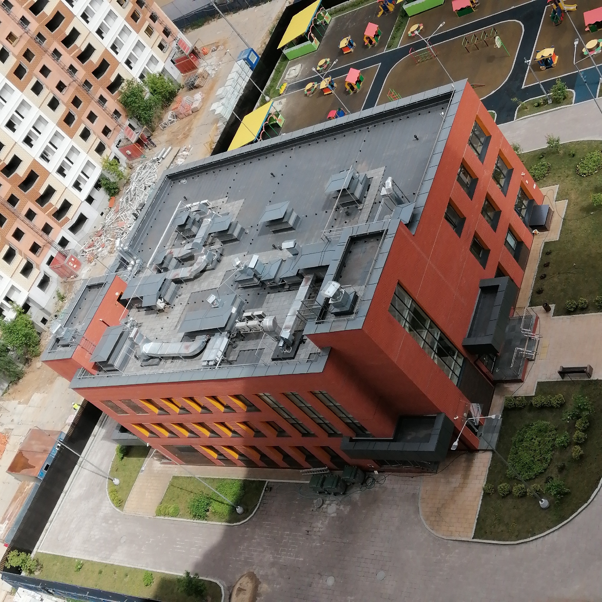 This aerial photograph captures an urban development featuring a completed modern building, an active construction site, and a children's playground.
The central subject is a contemporary building with a distinctive red-orange, textured facade and dark gray trim. Its flat roof is heavily outfitted with extensive metallic HVAC units, ducts, and other mechanical equipment, indicating a functional, climate-controlled interior. The building's windows are dark, some integrated with striking yellow or orange louvered sunshades on the lower levels. Several streetlights are positioned around the building, which is surrounded by green lawns, shrubs, paved walkways, and a small area with a bench. Large green electrical boxes or generators are situated near its base.
To the left, a tall residential building is clearly under construction, showing its exposed concrete framework and partially completed brick infill. Piles of construction materials, including bricks and metal debris, are scattered around its foundation, along with a blue portable toilet and other industrial containers.
Adjacent to the red-orange building, a vibrant children's playground features a dark, soft ground surface with lighter gray pathways. Numerous colorful play structures, such as slides, swings, and small sheltered areas with yellow and green canopies, are arranged across the area, suggesting a facility for children like a school or kindergarten.
The scene is illuminated by daylight, likely mid-day, under an overcast or partly cloudy sky, as indicated by the diffused lighting and soft shadows. No people or discernible text are visible in the image.