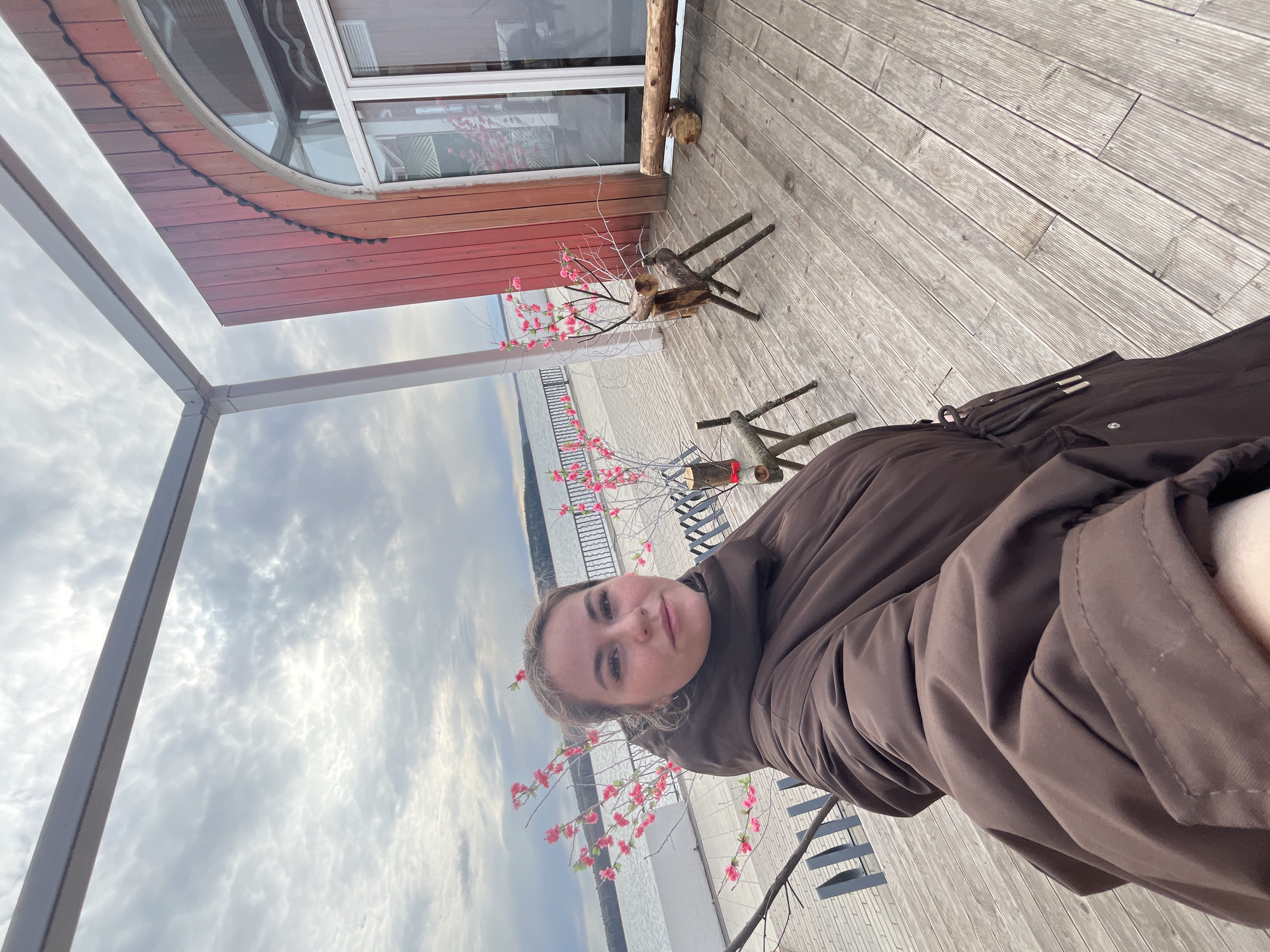 A photograph taken outdoors in the city of Chaikovsky, Russia, shows a woman from a high angle, likely a selfie. The woman has fair skin, light hair, and is wearing a brown jacket. The background includes a wooden deck, a body of water, and a cloudy sky. Artificial cherry blossom decorations in small wooden stands are visible along the deck's edge. A building structure with a rounded window is also present.