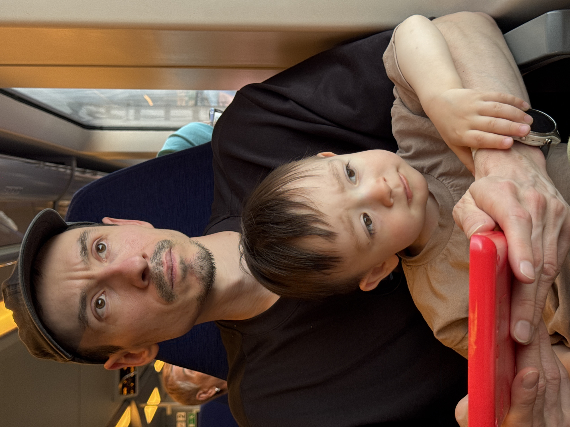A man wearing a gray cap and a black t-shirt is sitting with a young child on what appears to be a train. The child is leaning against the man. The train car is visible through the window in the upper left. Another passenger is visible in the reflection on the lower part of the window. The location is unknown and can not be determined from the information available.