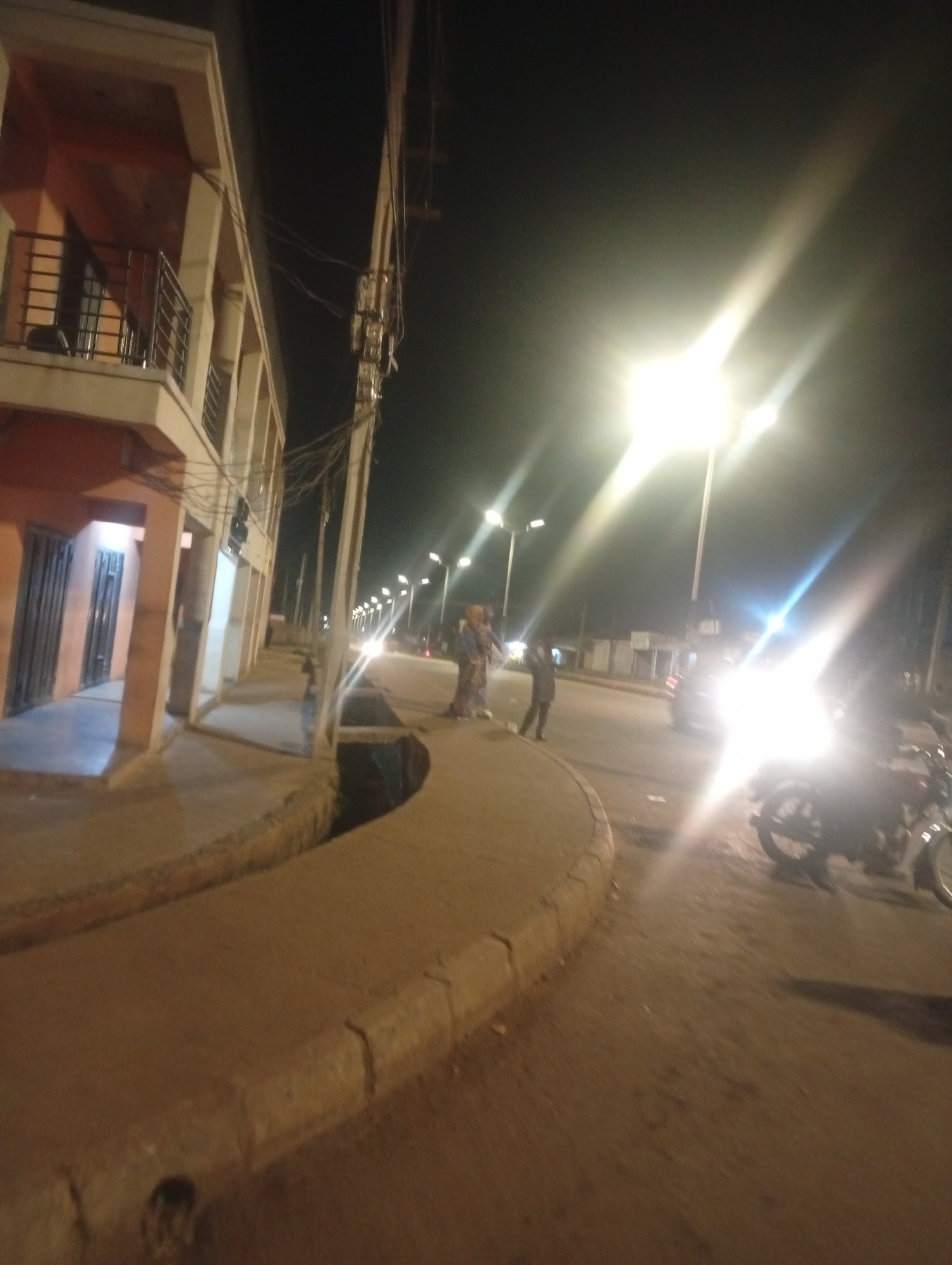 The image depicts a street scene at night in Bukuru, Nigeria, illuminated by streetlights and the headlights of a vehicle. On the left, a multi-story building with balconies and doors is visible. The street itself is paved, with a raised concrete walkway on the left, bordered by a drainage ditch. Several people are present, including a group gathered near the building on the left and individuals walking on the street. A motorcycle is parked on the right side of the road. The overall atmosphere is dimly lit, with strong light flares from the artificial sources, obscuring some details of the background and distant elements of the scene. The image appears to be captured from a low angle, looking down the street.
