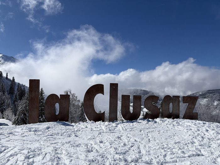 The image displays a panoramic view of a snowy alpine landscape in La Clusaz, France. In the immediate foreground, large, freestanding individual letters, constructed from a dark, weathered metal with a rusty appearance, spell out "La Clusaz." These letters are partially embedded in a thick layer of white snow, which shows varied textures from foot traffic or wind patterns.
Behind the prominent sign, dense evergreen forests, heavily laden with snow, cover the mid-ground mountain slopes. Further in the background, higher, snow-capped mountain peaks are visible, partially obscured by expansive white cumulus clouds. The sky above is a clear, vibrant blue. No human figures or active subjects are present within the scene.