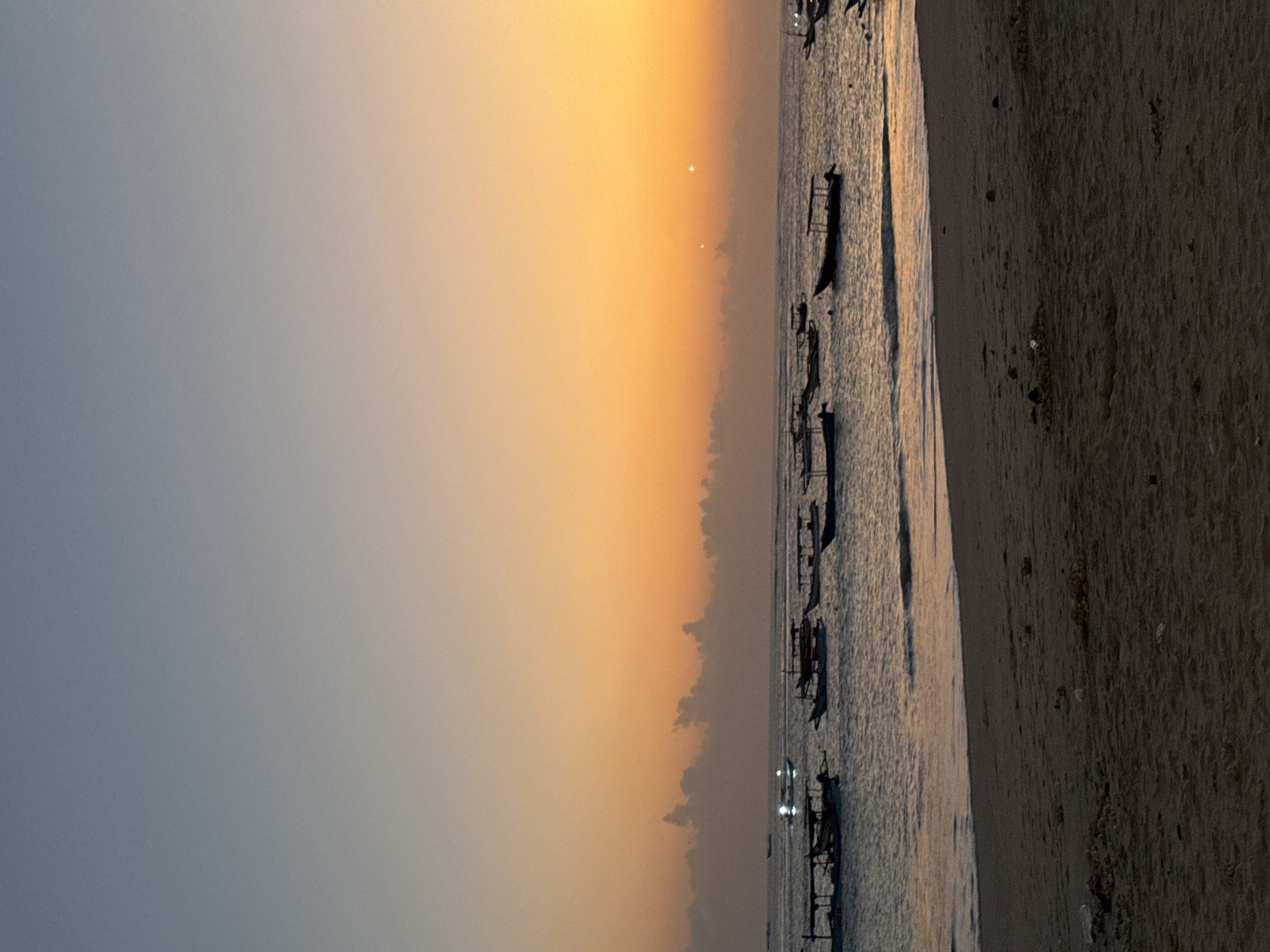 The image depicts a tranquil scene at Kuta Beach, Indonesia, during sunset. The sky is painted with a gradient of soft blues and warm oranges, transitioning into a bright yellow hue near the horizon. A few scattered clouds are visible, adding texture to the sky. The sea is calm, with gentle waves lapping at the shore. Numerous traditional Balinese outrigger boats, known as jukung, are anchored or gently floating on the water, their silhouettes dark against the colorful sky. Some of the boats appear to have canopies, suggesting they are used for leisure or fishing. The foreground is dominated by the sandy expanse of Kuta Beach, with some scattered debris and textures on the sand. The overall atmosphere is serene and peaceful, capturing the beauty of a tropical sunset.