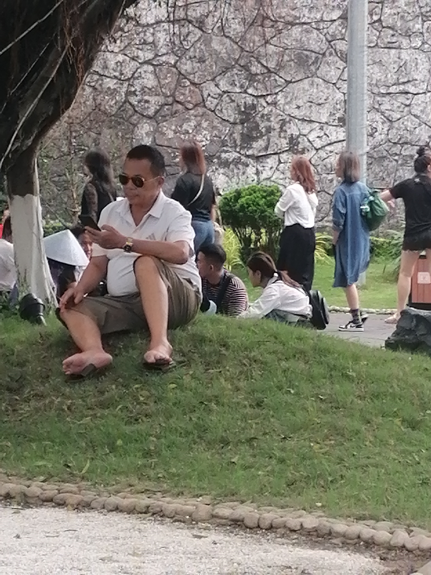 A middle-aged man wearing sunglasses, a light-colored collared shirt, and khaki shorts is seated on a grassy hill, holding a smartphone in his left hand. He is wearing flip-flops. Behind him, several people are present, including a woman in a white shirt and black skirt, a person in a blue dress, and others further away. A conical hat is visible next to the man. A gray stone wall is in the background, and a tall metal pole stands to the right of the frame. The location is Hong Gai Ward, Vietnam.