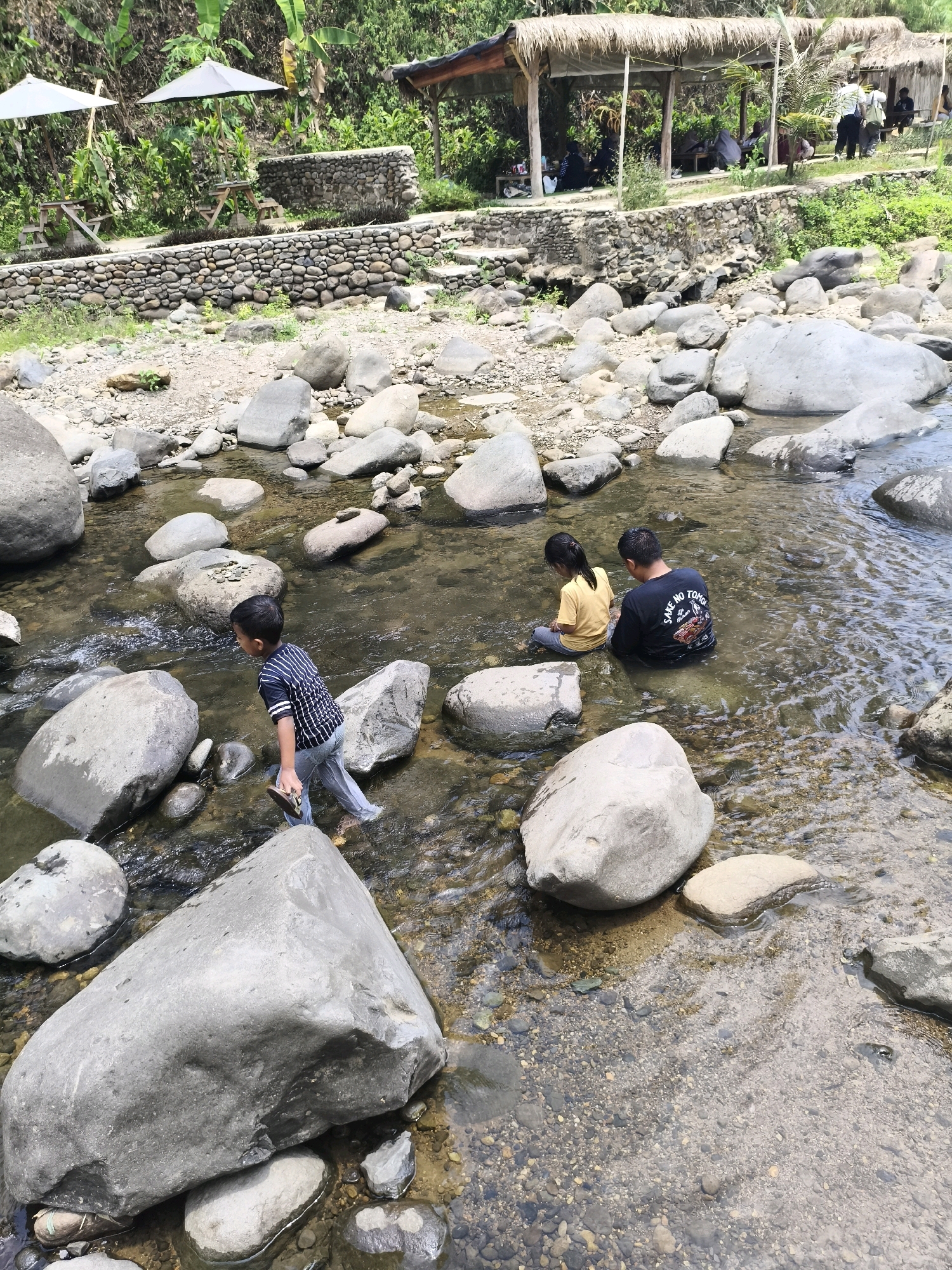 In the city of Babakan Madang, Indonesia, a shallow, rocky river flows through a lush green environment. In the foreground, three individuals are wading in the clear water. On the left, a young boy wearing a striped t-shirt and grey pants walks through the river, holding a flip-flop in his right hand. To his right, a young girl in a yellow t-shirt and grey pants sits in the water. Beside her, a man in a black t-shirt with a graphic print is also seated in the river. The riverbed is visible, covered with pebbles and larger stones. In the background, a stone wall separates the river from a raised area with picnic tables and umbrellas, suggesting a recreational spot. Beyond that, a thatched-roof pavilion with people gathered under it is nestled amongst dense vegetation. The overall scene depicts a relaxed outdoor activity in a natural setting.