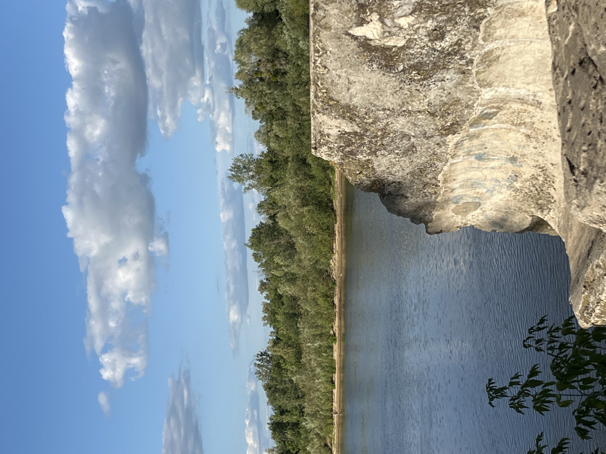 The image depicts a serene natural landscape with a wide body of water in the foreground. The water is calm, with gentle ripples on its surface. To the right, a large, textured rock formation juts out into the scene, its rough surface hinting at age and exposure to the elements.
Behind the water, a dense line of trees forms a lush green backdrop. The foliage is varied, suggesting a mix of deciduous and possibly coniferous trees. A sandy bank is visible between the water and the treeline, indicating a riverbank or shoreline. The sky above is a clear blue, dotted with scattered white clouds, some of which are fluffy and cumulus-like, while others are more wispy. The overall impression is one of a peaceful, undeveloped natural environment.
The location is the city of Narowlya, Belarus, likely along the Pripyat River, which flows through the region. The presence of the river, the natural vegetation, and the absence of urban development strongly suggest this setting. There is a single, faint silhouette of a person visible in the distance along the riverbank, barely discernible against the treeline. The focus of the image is the natural scenery, with the rock formation in the foreground adding a sense of scale and texture to the composition.