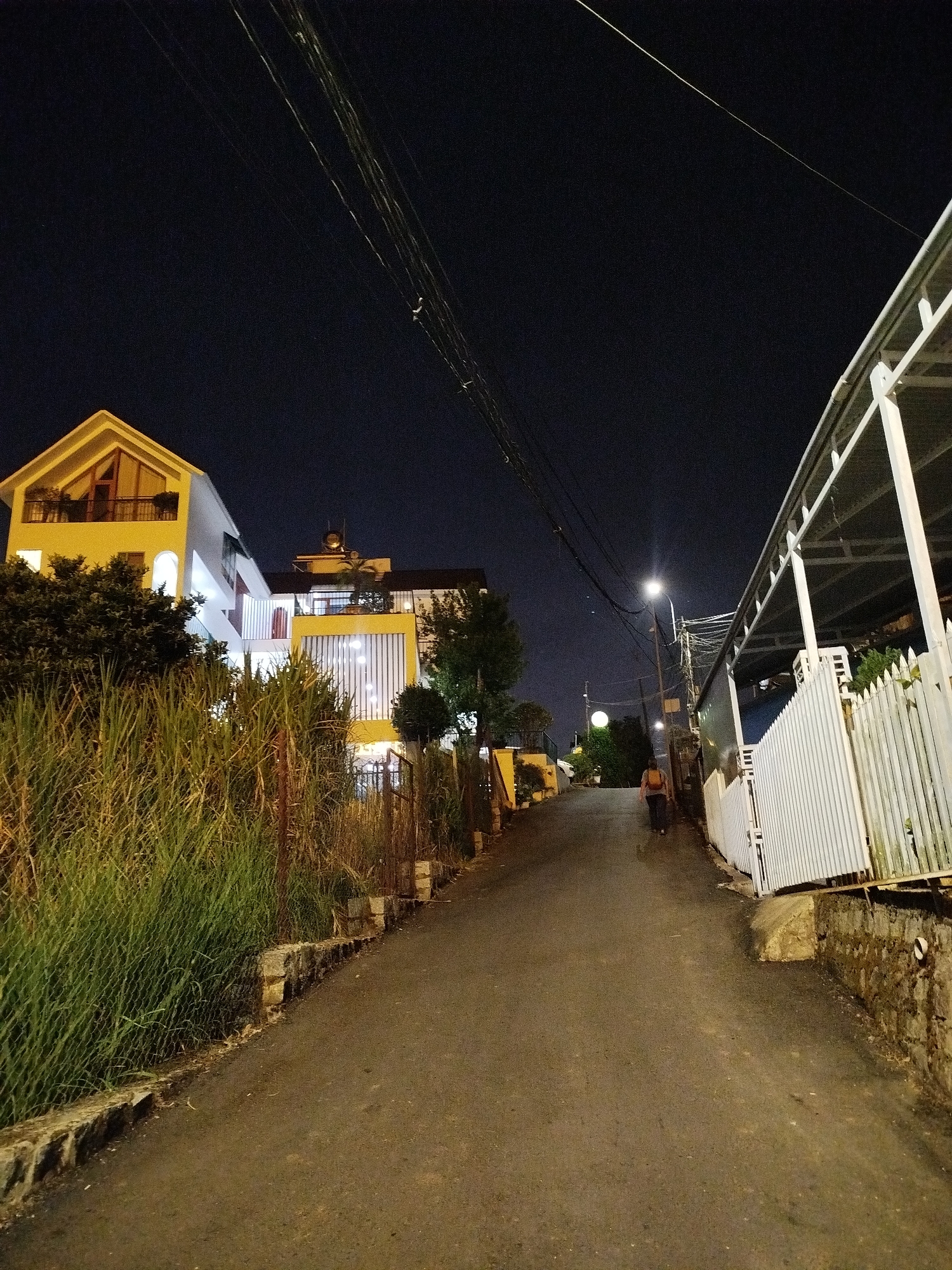 A street scene at night in Da Lat, Vietnam. The asphalt road curves uphill and away from the viewer. A figure wearing a backpack is walking away from the viewer further up the road. Overhead electrical wires are visible against the night sky. A light-colored building is located on the left of the street, surrounded by dense vegetation and fencing. The building appears to be a residence with visible interior lighting. On the right side of the street, a white fence and roof structure are present. The overall light level is low, and the scene is illuminated by streetlights and building lights.