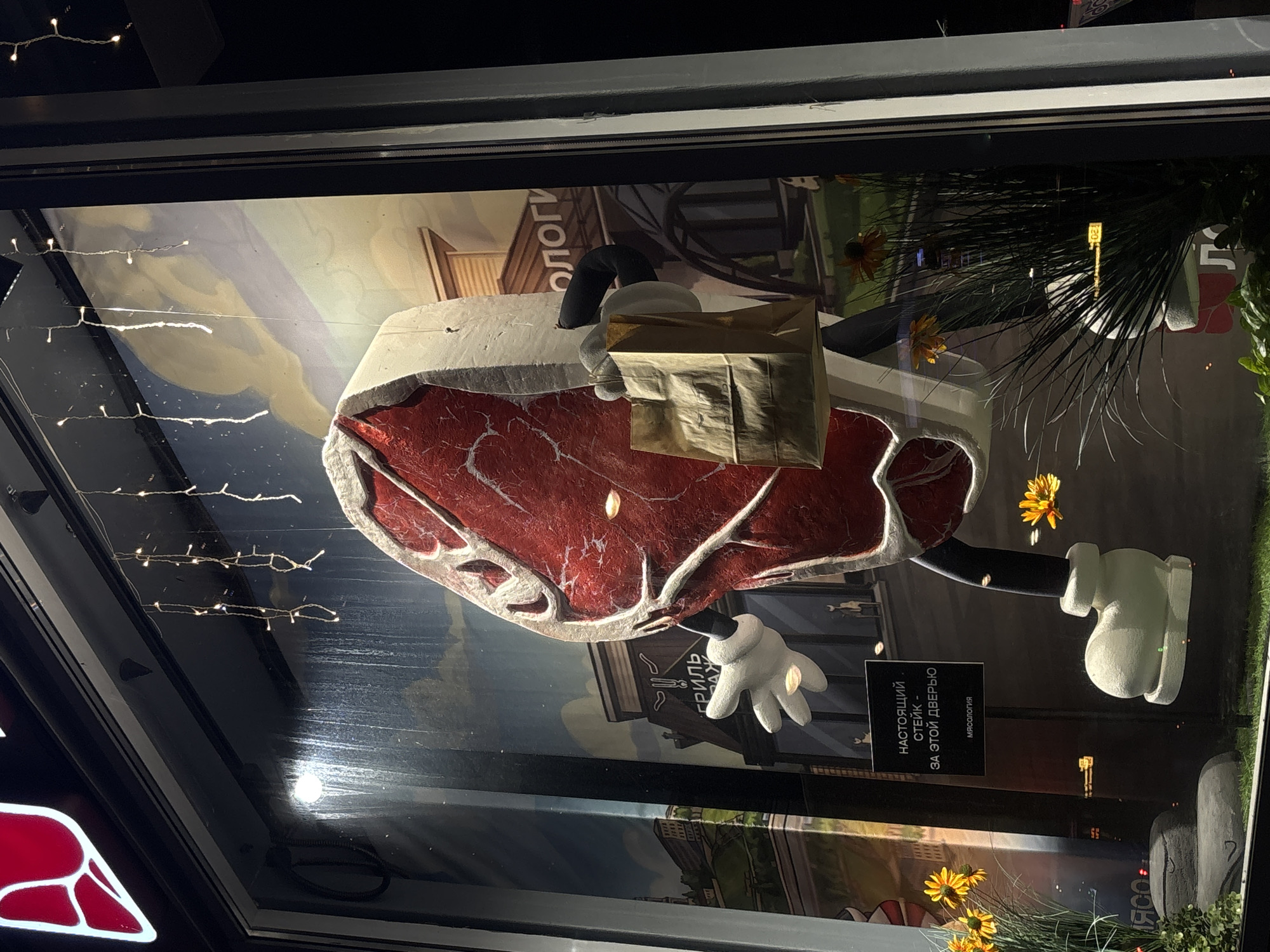 In a storefront window in Perm, Russia, a whimsical display features a large, anthropomorphic steak character holding a paper bag. The steak, with a marbled red interior and white fat edges, is depicted with cartoonish white gloves and oversized white shoes. It stands in front of a painted mural that resembles a street scene with buildings and a sky with clouds. String lights hang in the window, adding a festive atmosphere. A sign on the display reads "НАСТОЯЩИЙ СТЕЙК - ЗА ЭТОЙ ДВЕРЬЮ" which translates to "REAL STEAK - BEHIND THIS DOOR" and "МЯСОЛОГИЯ" which translates to "MEATOLOGY." The display is further enhanced by artificial plants and flowers, creating an eye-catching advertisement for the establishment.