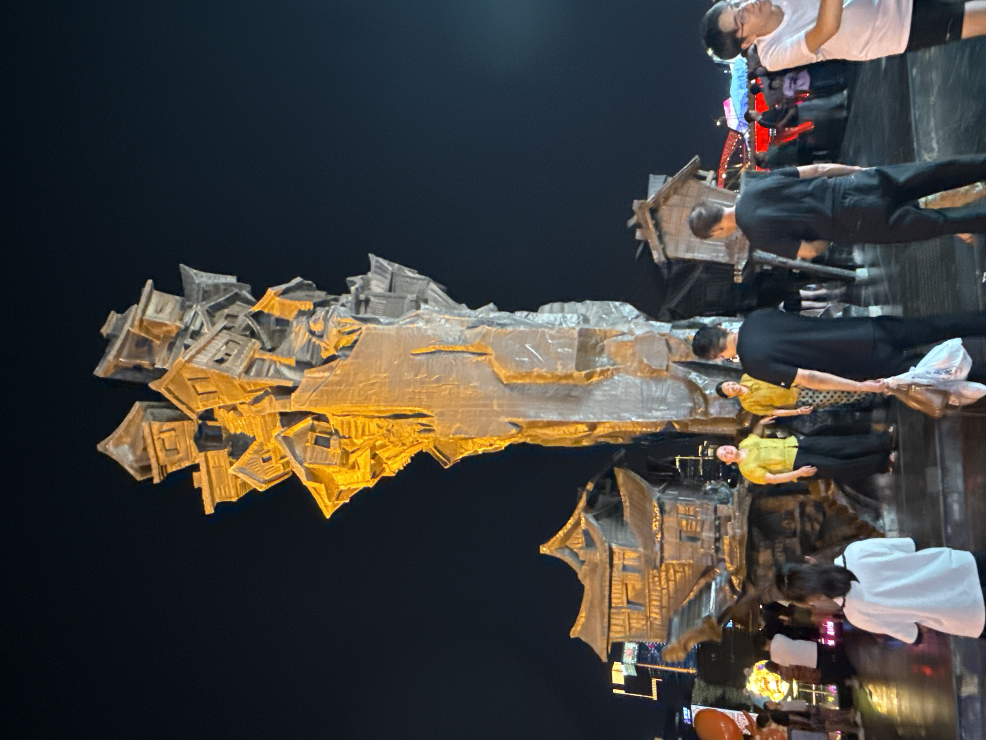The image depicts a nighttime scene in the Yuzhong District of China, featuring a large, abstract sculpture composed of stacked, stylized houses and a central, rough-hewn column. The sculpture is illuminated with warm yellow light, creating a dramatic effect against the dark sky. Several people are visible in the foreground and midground, interacting with the environment.
In the immediate foreground, a person wearing a white shirt with black trim faces away from the camera, their back to the viewer. To their right, a person in a black t-shirt and black pants walks towards the right, carrying two plastic bags. Further to the right, a person in a white t-shirt and dark shorts with glasses is visible from the side.
In the midground, two women stand near the sculpture. The woman on the left wears a bright yellow short-sleeved shirt and black pants. The woman on the right wears a similar yellow shirt and patterned pants. Behind them, more people are seen in the background, some walking and others gathered. The sculpture itself is intricate, with many small, angular house-like structures clustered at the top of the central column, resembling a chaotic, upward growth. Below the main sculpture, there are smaller, more distinct house structures integrated into the base. The ground appears to be wet, reflecting the lights from the surroundings. In the far background, city lights and structures are visible, suggesting an urban setting. The overall atmosphere is that of a public gathering or attraction at night.