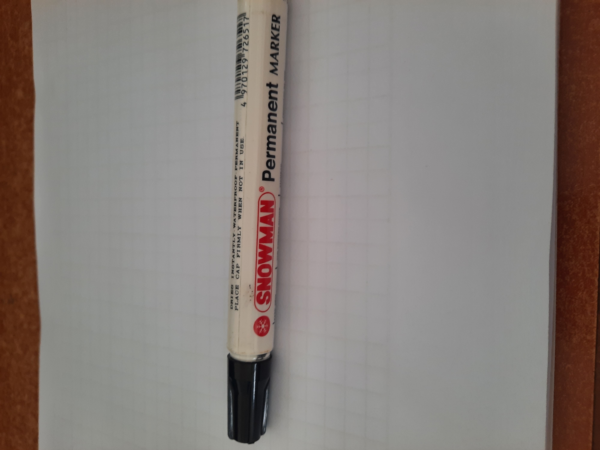 A black-capped, white permanent marker with the brand "SNOWMAN" and the words "Permanent MARKER" printed on its side is positioned horizontally on a white, grid-lined paper. The marker is slightly angled, with the cap on the left side and the tip facing right. The background consists of a wooden surface above and below the paper. The image does not provide any information about the city of Lhokseumawe, Indonesia.