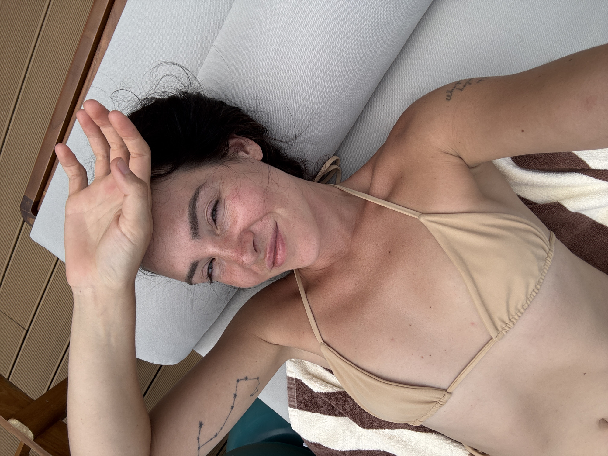 A woman is lying on a lounge chair outdoors, possibly on a deck, in North Kuta, Indonesia. She is wearing a tan bikini top and has her left arm raised, shielding her eyes with her hand. Her hair is dark and appears slightly windblown. The lounge chair is light gray, and a striped towel is partially visible. The background shows a wooden deck and a glimpse of a railing. There is a tattoo of a constellation on her left arm.