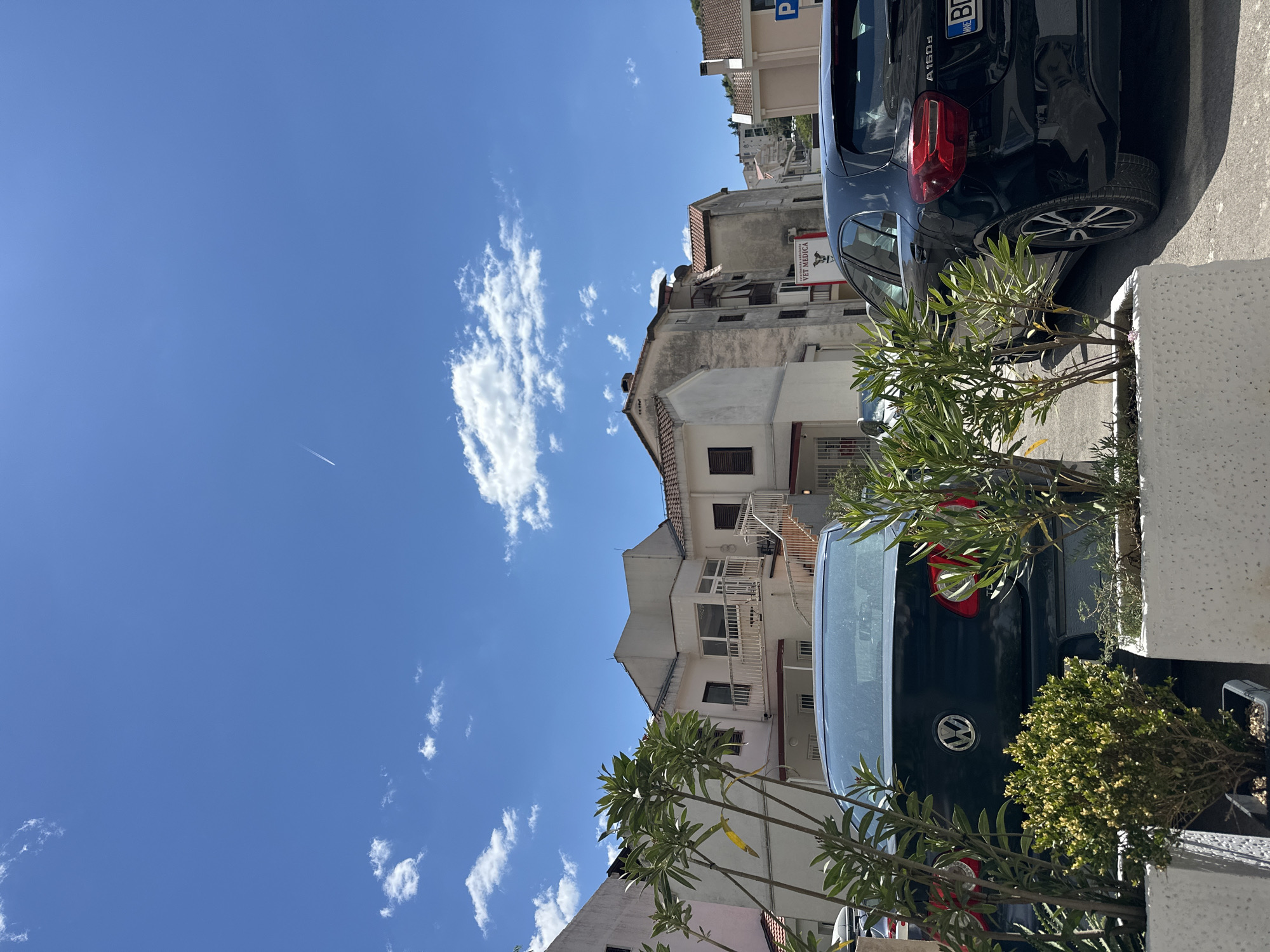 The image shows a street scene in Budva, Montenegro on a sunny day. Two black cars are parked in a row, with a planter in the foreground containing green plants. Several buildings are visible, including a tall, older building with multiple floors and windows. The sky is blue with scattered white clouds and a contrail from an airplane. The buildings are faded, and have aged, but are also maintained. A sign is also visible on one of the buildings.