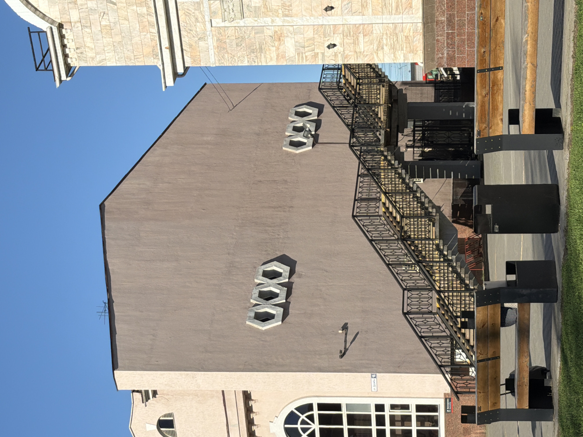 A building with a light brown exterior is positioned under a clear, blue sky. The architectural style of the building includes a set of three hexagon-shaped, decorative window frames on its face and a black metal fire escape stairwell. A wooden structure and a black, box-shaped trash receptacle are positioned near the building's base along a sidewalk. The building's location is indicated by a small street sign, which appears to read "ул. Дружбы, 16," suggesting a location in Izhevsk, Russia. A portion of an adjacent building with light-colored brickwork is visible in the background.