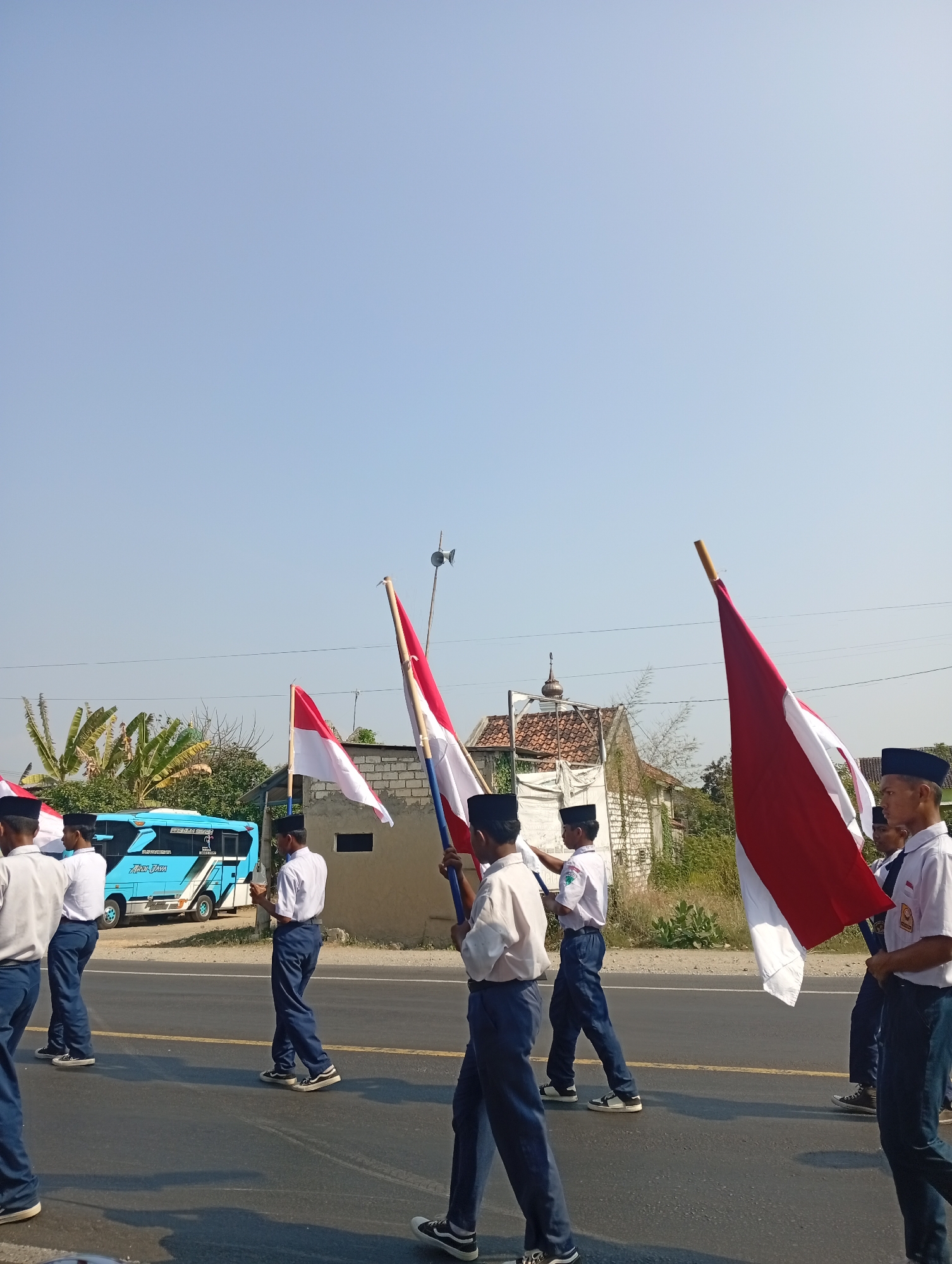 In the city of Sarang, Indonesia, a group of students in uniform are participating in a parade. They are marching on a paved road under a clear blue sky. Each student is holding a flag, with the Indonesian flag, red and white, being the most prominent. In the background, a bright blue bus with "Putra Jaya" written on its side is visible, along with some residential buildings and lush green vegetation. The students appear to be in organized formation, suggesting a formal event or ceremony.
