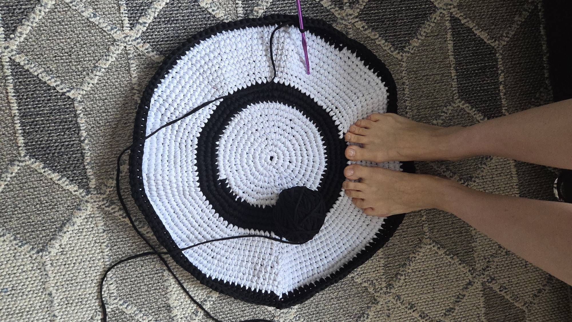 The image captures an overhead view of a crafting project in a home setting, inferred to be in Chaikovsky, Russia. The central subject is a large, circular crocheted mat with a bold black and white concentric ring pattern. The mat starts with a white center, surrounded by a black ring, then a wider white ring, and an outermost black border that is currently being worked on. The stitches are large and appear to be made from thick fabric strips or chunky yarn.
A purple-handled crochet hook is visible, inserted into the white section of the mat, indicating the project is actively being worked on. A small ball of black yarn rests on the mat near the center, with a strand extending to the crocheted piece.
A pair of bare feet, presumably belonging to the craftsperson, are positioned on the outer white ring of the mat. The feet are relaxed, with natural-colored toenails. The mat is laid on a textured rug featuring a geometric pattern in various shades of grey, beige, and darker tones, providing a cozy domestic backdrop. The scene suggests a calm, creative activity taking place indoors. No visible text is present in the image.