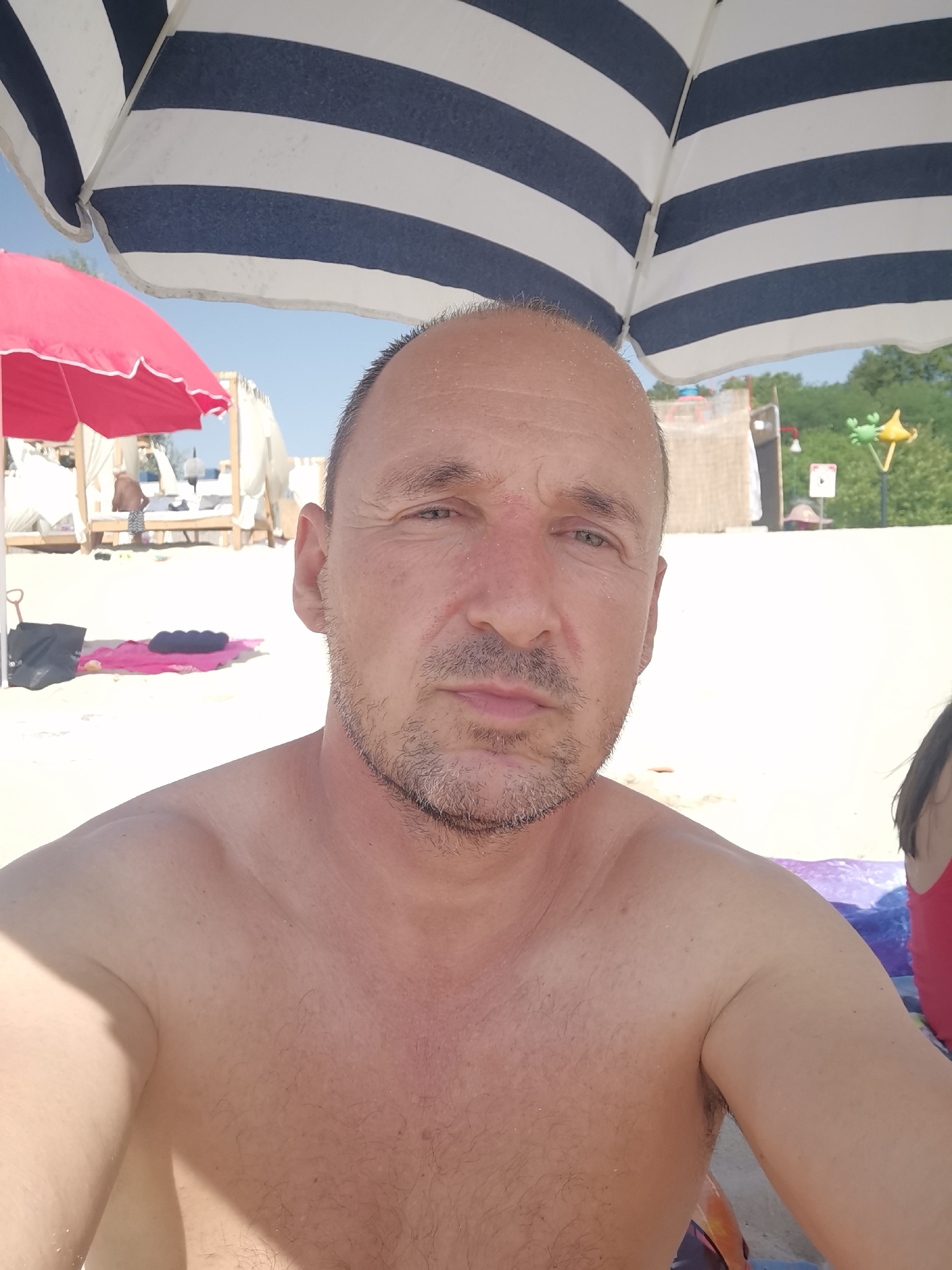 A man is taking a selfie on a beach in Kranevo, Bulgaria. He is bald with stubble on his chin and is shirtless, showing his shoulders and chest. He is sitting under a large blue and white striped umbrella. In the background, there is a pink umbrella, beach huts with white drapes, and trees. There are also beach towels and bags on the sand. The man appears to be looking at the camera with a neutral expression. The lighting suggests it is a sunny day.