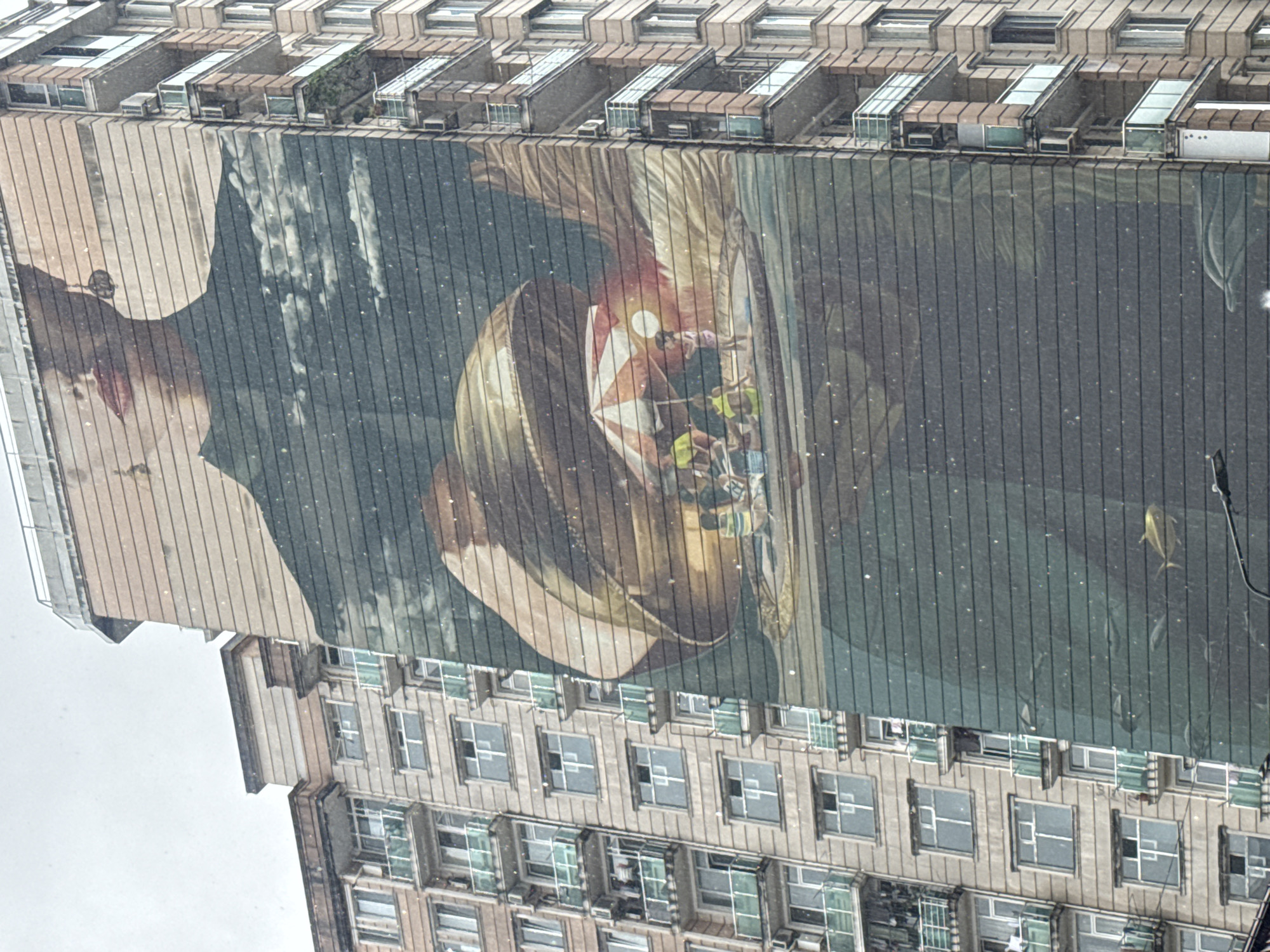 A large building with multiple floors has a very large mural covering a large portion of its exterior wall. The mural depicts a man's face and a bowl with people in it. The building is in Batumi, Georgia.