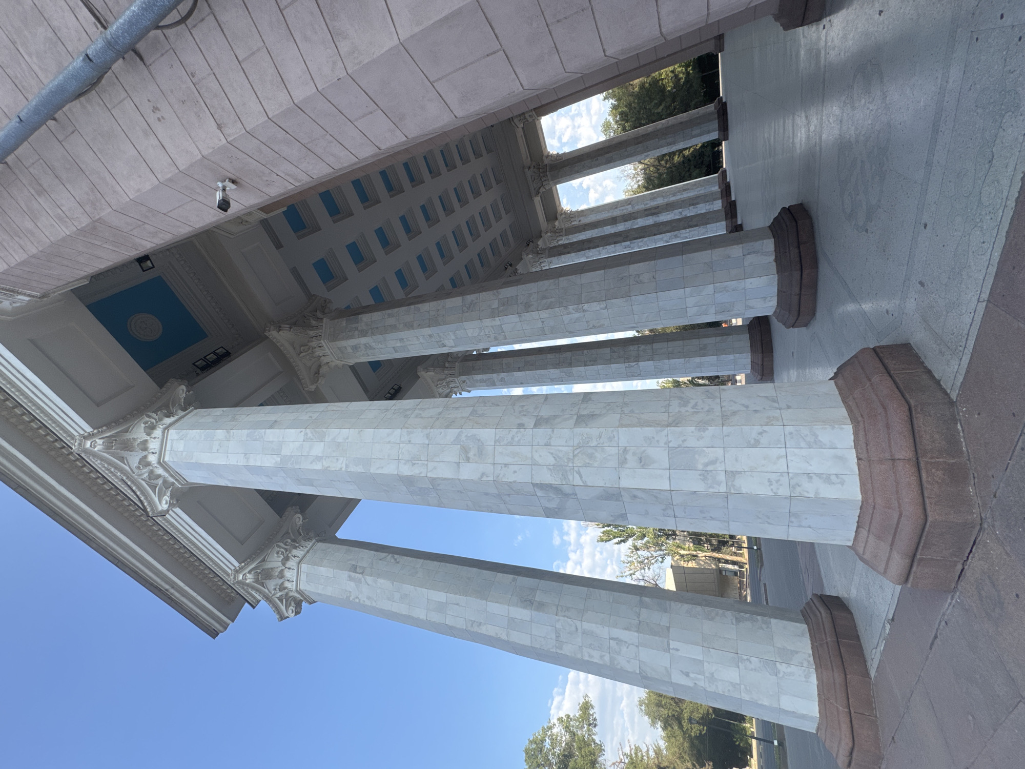 The image shows a wide-angle view of the colonnade of the State Historical Museum in Bishkek, Kyrgyzstan. The museum's facade features a series of imposing, white marble-clad columns with Corinthian capitals. The columns are set against a clear blue sky with scattered clouds. The camera is positioned low, looking upwards, emphasizing the grandeur of the architecture. The ground in front of the colonnade is paved with light-colored stone tiles. The ceiling of the colonnade is visible, with blue recessed panels and intricate molding. A security camera is mounted on the exterior wall to the right. Trees and other buildings are visible in the background through the spaces between the columns.
