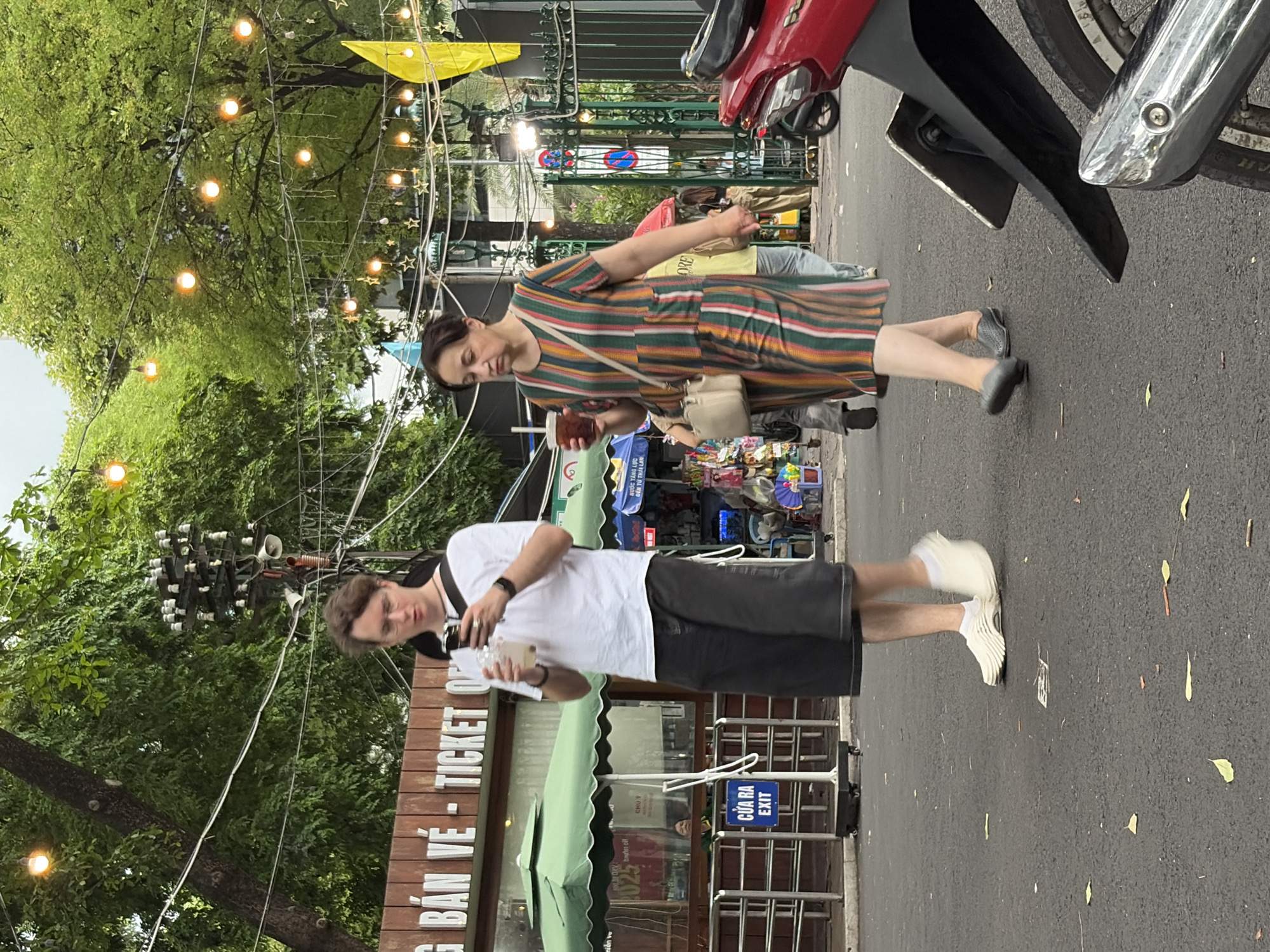 This image captures an outdoor urban scene in Thủ Đức, Vietnam, featuring two pedestrians and background elements suggesting a ticketed area or attraction.
In the foreground, an asphalt surface with scattered leaves and small debris is visible. On the left, a young adult male with light skin, short brown hair, and glasses walks towards the right. He is wearing a white short-sleeved t-shirt, dark wide-leg shorts, white socks, and light-colored slip-on shoes. He holds a clear plastic cup with a light beverage in his left hand and is looking down at a smartphone in his right hand. A black strap is around his neck.
On the right, a middle-aged to older adult female with light skin and dark hair pulled back walks towards the left. She is wearing a vertical striped midi-dress in green, orange, white, and a darker hue, along with dark grey slip-on shoes. She carries a light beige shoulder bag with a long strap and holds a clear plastic cup containing a dark beverage in her right hand.
The two individuals are walking in opposite directions and do not appear to be interacting.
In the midground on the left, a brown wooden sign with white lettering reads "G BÁN VÉ - TICKET OF" (partially visible), indicating a ticket office. Below this, a green umbrella or awning is open. A metal barrier system is present, behind which a blue sign with white text reads "CỬA RA EXIT." A person's face is visible inside a structure behind the barrier, likely a ticket booth window.
On the right midground, another green umbrella/awning covers a stall displaying various colorful merchandise. Further back, an ornate dark green metal gate is visible. Part of a red motorcycle, including its rear fender, exhaust pipe, and tire, is visible on the far right.
The background is dominated by lush green trees with dense foliage. Numerous strings of warm yellow globe lights and star-shaped decorative lights are strung across the scene between the trees. A utility pole with multiple white ceramic insulators and a loudspeaker is visible behind the male pedestrian. A bright yellow flag or banner hangs from a pole on the far right of the background. The sky appears overcast, and the string lights are illuminated, suggesting either evening or a purely decorative function during the day.