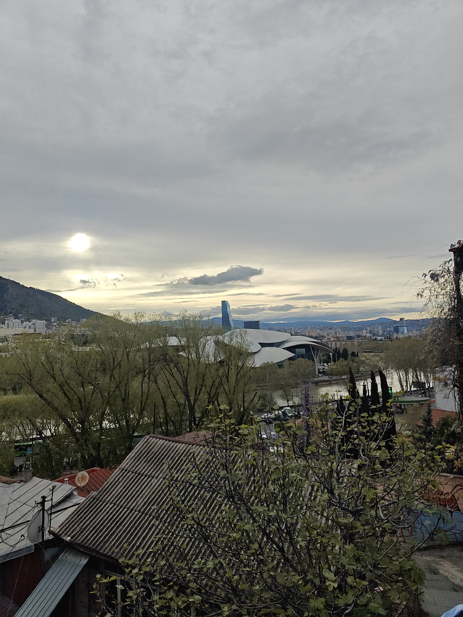 An elevated city view of Tbilisi, Georgia, captures a blend of old and new urban landscapes under a cloudy sky with the sun breaking through.
In the immediate foreground, weathered rooftops with corrugated metal and tile patterns are visible, along with a satellite dish and the bare branches of a bushy tree showing nascent green leaves. Further down, another tree, larger and denser with green foliage, partially obscures a retaining wall with faded, undecipherable graffiti.
The midground features the Mtkvari River flowing through the city, flanked by numerous trees, some with early spring leaves. Along the riverbanks, modern architectural landmarks stand out, including the distinctive, shell-like structures of what appear to be the Rike Park Concert Hall and the undulating glass and steel of the Public Service Hall. A road with parked cars is also visible near the river.
The background reveals the sprawling city of Tbilisi, extending across valleys and up distant hills. On the far left, a prominent, verdant mountain or large hill (likely Mtatsminda) rises, dotted with buildings. In the distance, layers of mountains frame the horizon. The sky is largely overcast with scattered clouds, but the sun is visible low on the left, creating a bright patch of light that suggests either early morning or late afternoon.
The overall weather is cloudy, providing diffused light. No people are discernible in the scene. The visible text on the graffiti wall is not legible. The combination of historic rooftops, modern architecture, the Mtkvari River, and the surrounding mountains strongly places the scene in Tbilisi.