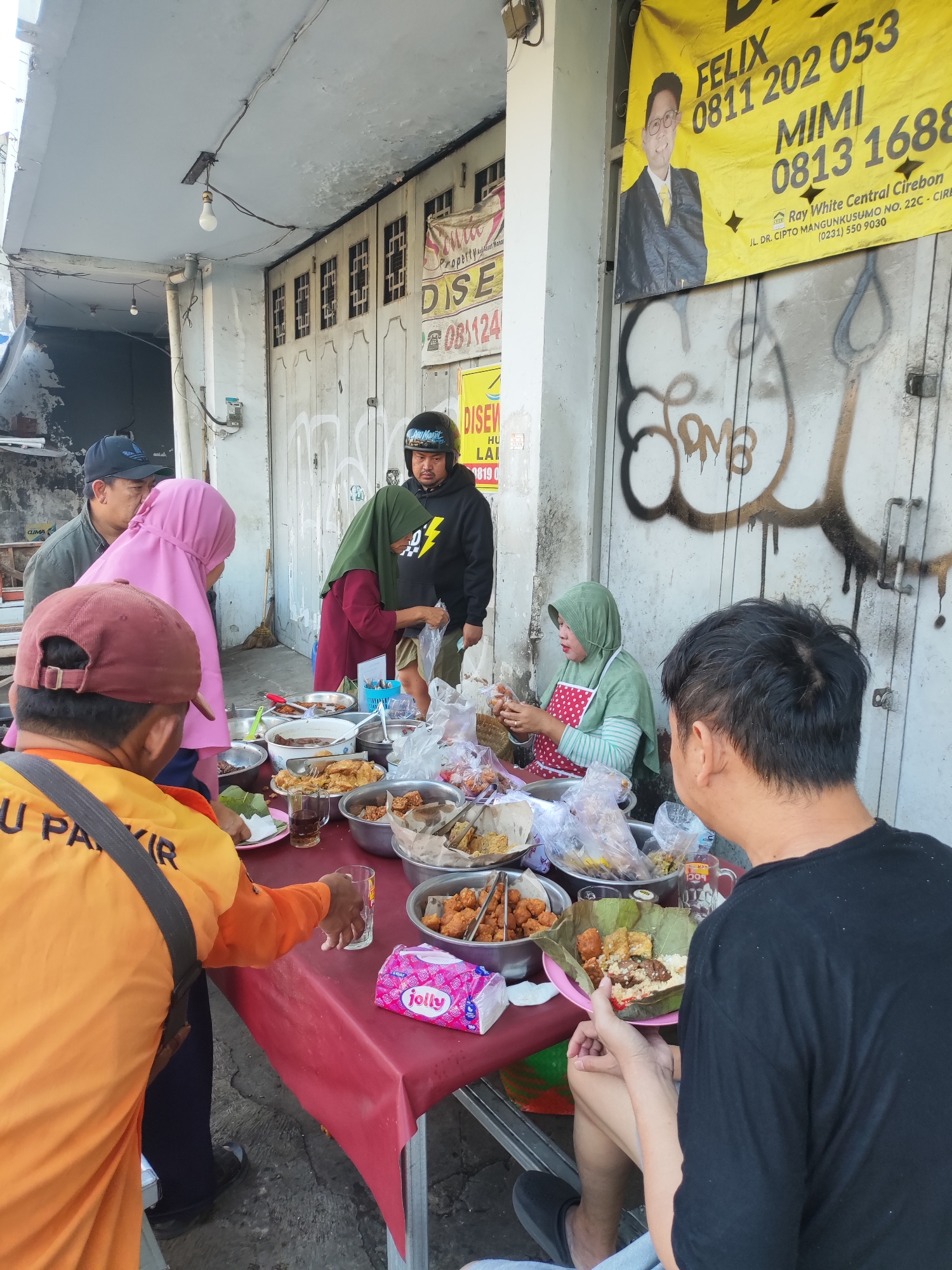 A group of people are gathered around a table laden with food, appearing to be enjoying a meal outdoors. The setting is an urban environment, possibly a street or sidewalk, with a building facade in the background featuring large metal doors with some graffiti. A yellow banner with text and an image of a man in a suit is prominently displayed on the building. The table is covered with a red tablecloth and holds various dishes, bowls of food, drinks, and a packet of tissues. Several individuals are in the process of serving themselves or eating. One person, wearing an orange jacket with "PARKIR" (parking) written on it, is reaching for a glass. Another person in a black t-shirt is holding a plate of food. A woman wearing a green headscarf and a red and white polka dot apron is serving food. Two other individuals, one in a pink hijab and another in a black hooded sweatshirt, are also near the table. The overall impression is of a casual, communal meal being shared in a busy street-side location. The presence of the yellow banner with contact information (phone numbers 0811 202 053, 0813 1688) and an address on "JL. DR. CIPTO MANGUNKUSUMO NO. 22C, CIREBON" confirms the location is indeed Cirebon, Indonesia.