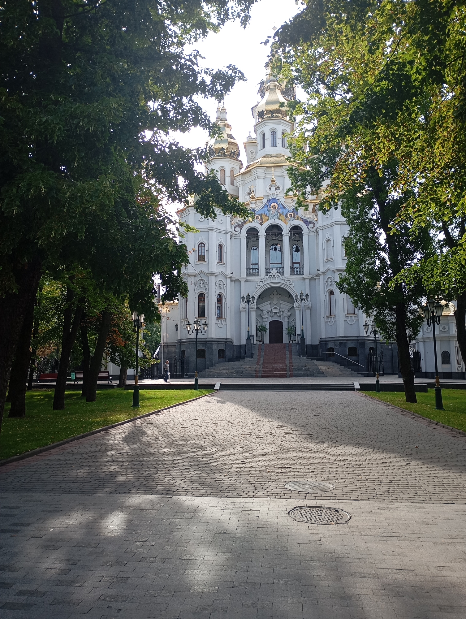 This image shows the Annunciation Cathedral in Kharkiv, Ukraine. The cathedral is a large, white building with golden domes, seen from the front through a line of trees. A paved walkway leads to the cathedral's entrance, which has a grand staircase. The walkway is bordered by a grassy area with trees and lampposts. The sun is shining, casting shadows on the walkway. There are a few people visible in the background, walking on the grass and near the cathedral. The scene appears to be set in a park or public square.