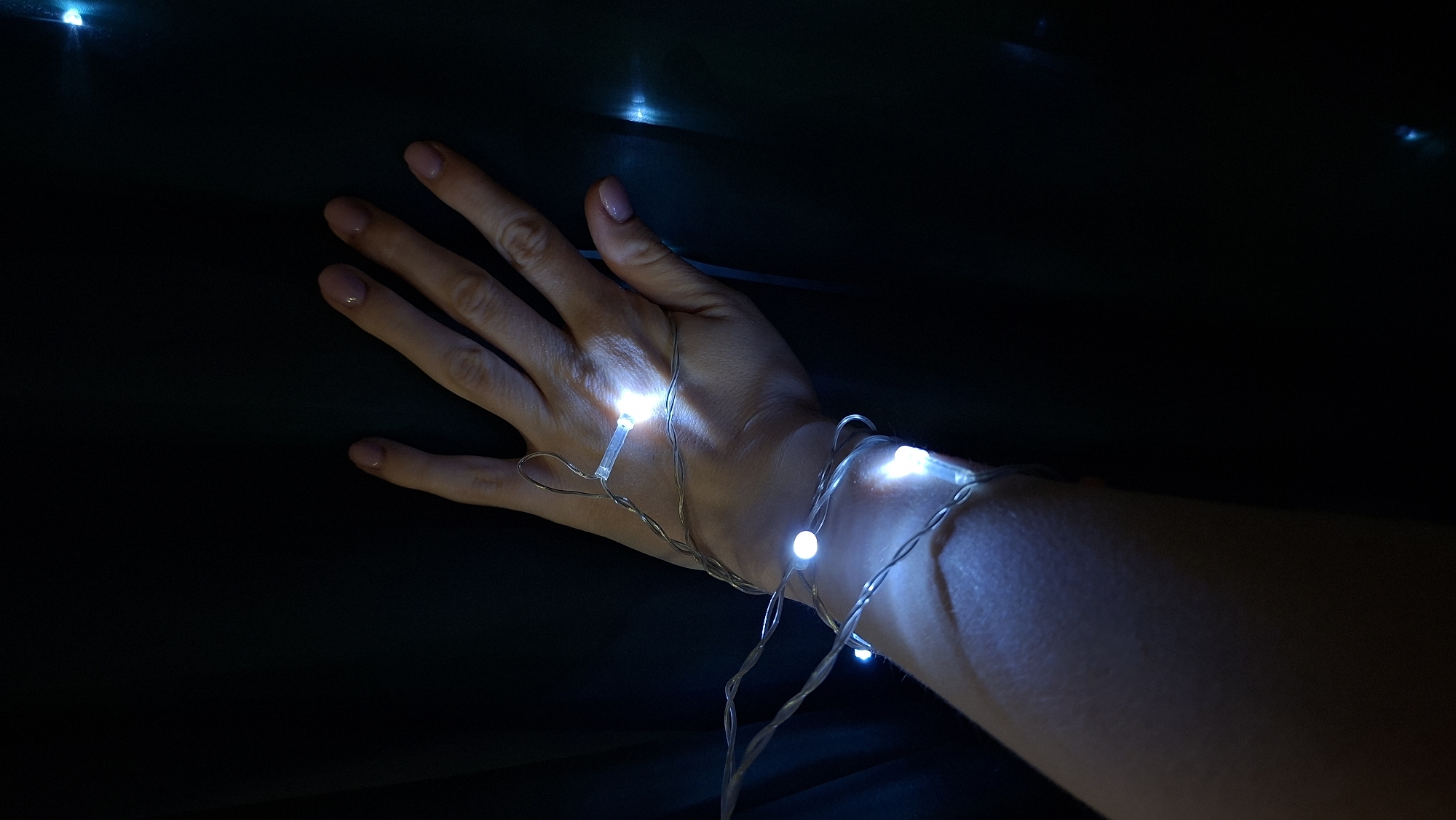 A Caucasian person's left hand and lower forearm are illuminated by small, cool-white LED string lights wrapped around the wrist and extending onto the back of the hand. The hand is open, with fingers slightly spread. The background is dark and mostly out of focus. The location in the city of Chaikovsky, Russia is not discernable from the image.