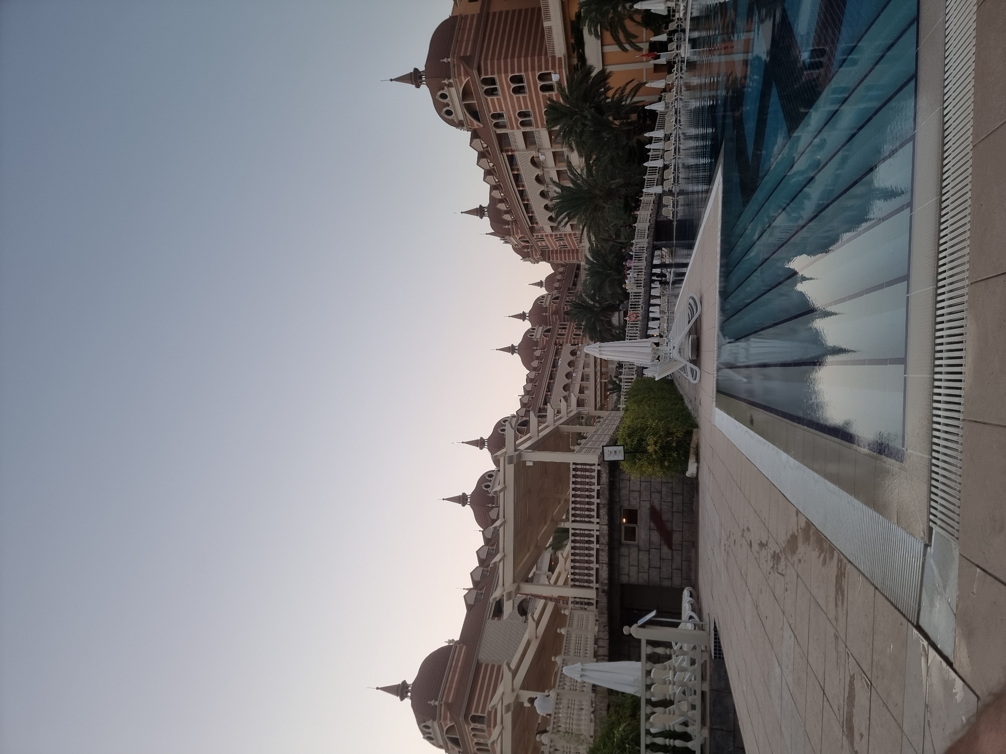 This is a photograph of the Side Crown Palace Hotel in Manavgat, Turkey, taken at dusk. The hotel is a large, multi-story building with a distinctive architectural style featuring red-tiled domed roofs, arched windows, and striped facades. In the foreground, a swimming pool with blue tiled steps is visible, reflecting the hotel and the sky. Lounge chairs and umbrellas are arranged around the pool area, and palm trees are interspersed among the hotel buildings. The sky is clear and pale, indicating the end of the day. A few people are visible on a bridge in the middle ground, and another person stands on a balcony on the left side of the frame. The overall scene suggests a tranquil resort atmosphere.