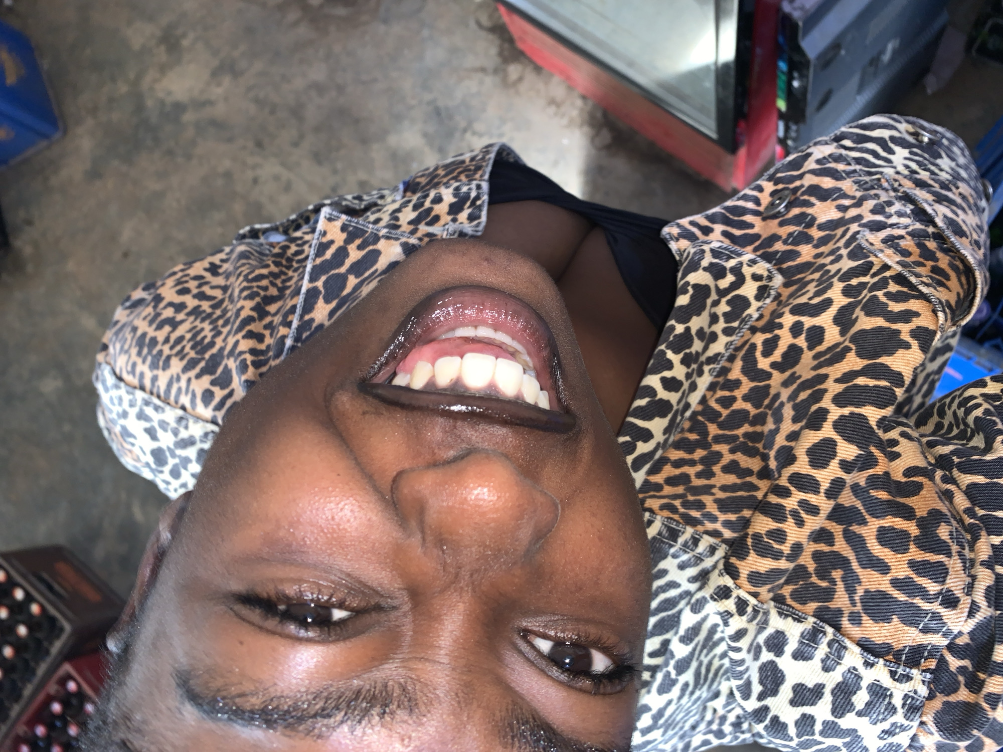 A young woman is captured in a close-up, head-and-shoulders shot, smiling broadly with her mouth open and teeth visible. She is wearing a leopard-print jacket over a black garment. The image is taken from a high angle, looking down at her. The background is blurred and appears to be an outdoor or semi-outdoor setting with some crates of bottles visible on the left and a red object that could be a vehicle or furniture on the right. The environment suggests a casual setting, possibly a market or a gathering place in New Stadium, Nigeria.