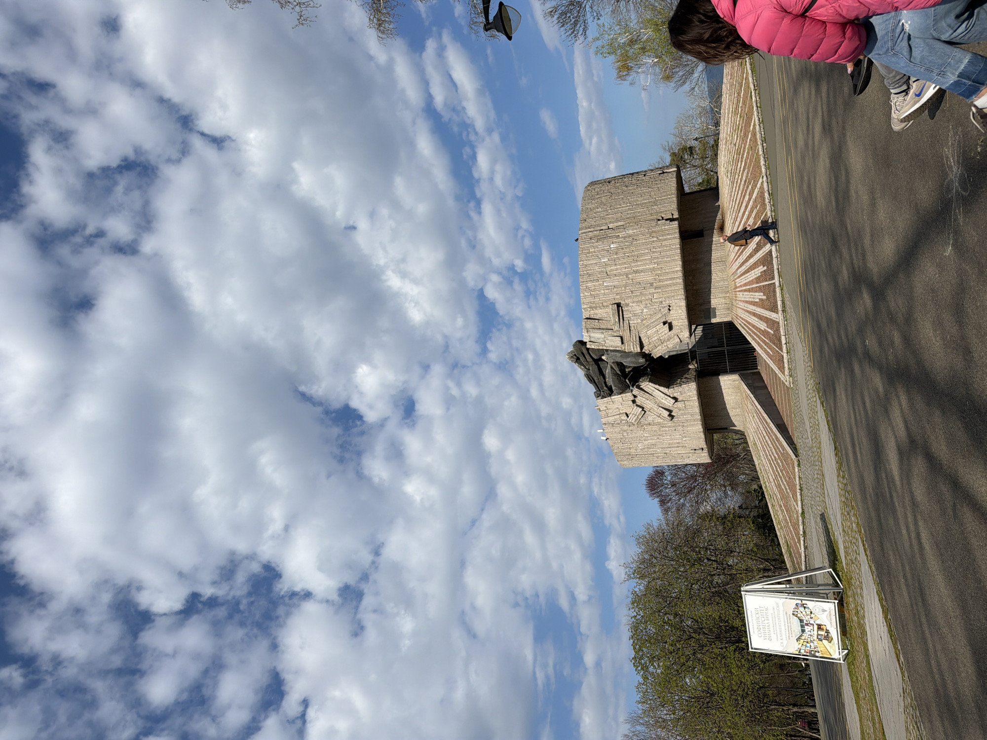 The image shows a monument and a cloudy sky. The monument appears to be made of stone. It is located in Burgas, Bulgaria. A person in a pink jacket is visible on the right side of the frame. There is a sign near a road with trees.