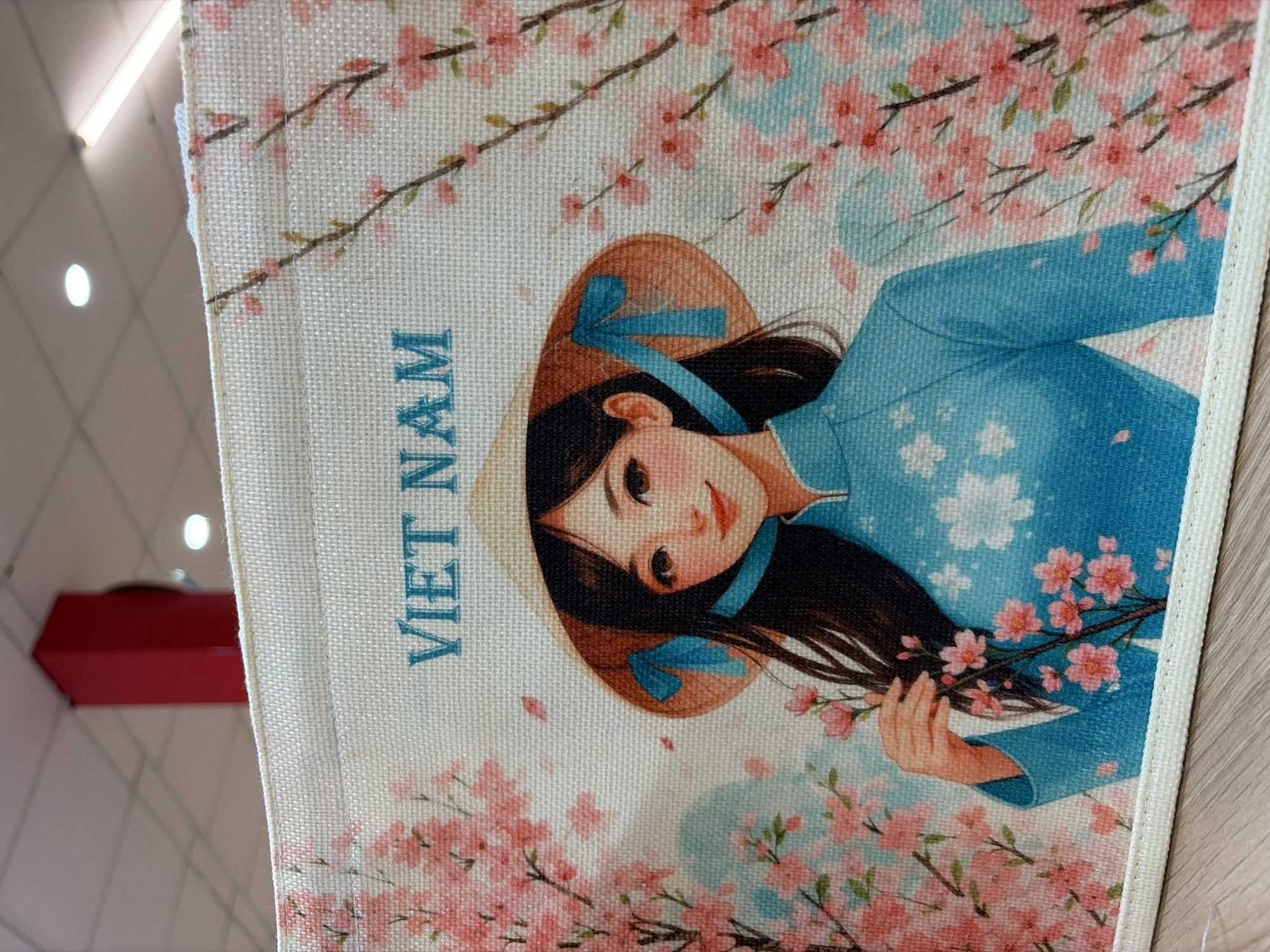 The image shows a close-up of a fabric item featuring a stylized illustration of a young woman wearing a conical hat and a traditional Vietnamese áo dài. The woman has long, dark hair and is holding a sprig of cherry blossoms. The background is adorned with more cherry blossom branches. The text "VIET NAM" is printed in blue capital letters above the woman's head. The fabric appears to be part of a storage box or decorative textile. The overall scene is depicted with soft, pastel colors. The image does not provide any information about the city of Đà Nẵng, Vietnam, or any specific location within it.