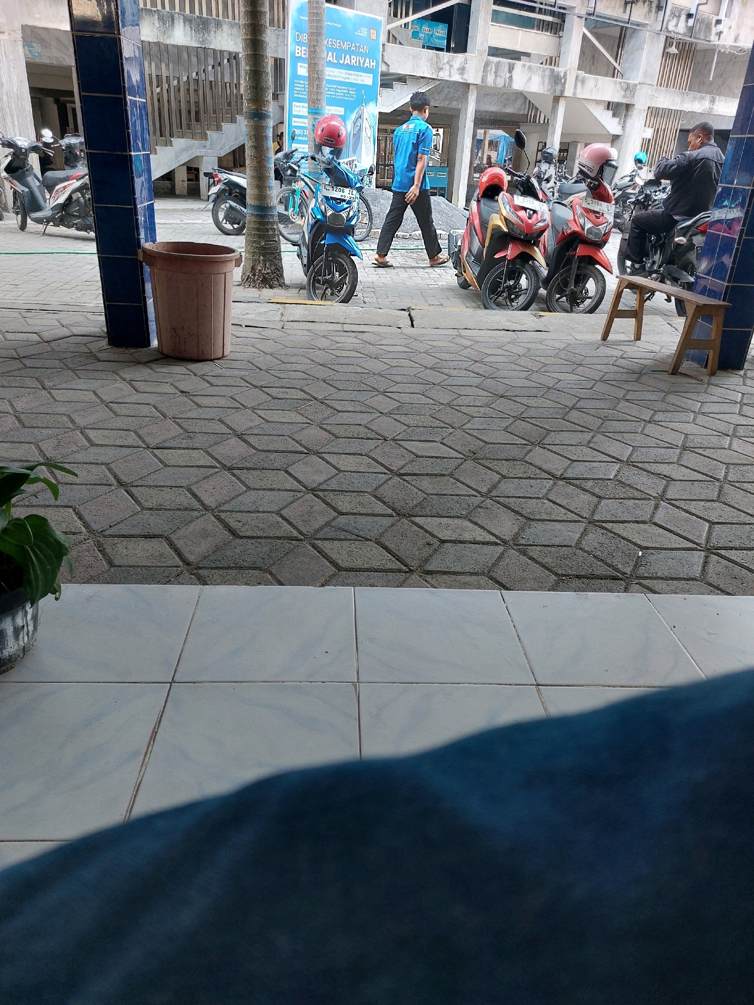 The image depicts an outdoor scene with a paved area in the foreground, composed of hexagonal-patterned interlocking stone tiles. Beyond this, a sidewalk of white square tiles leads to a paved walkway. A variety of parked motorcycles are visible, some with helmets on their seats. In the background, a building with concrete pillars and staircases is present, alongside palm trees and a large banner with Indonesian text. People are seen walking and sitting. One person in a blue shirt walks past the motorcycles, and another in a dark shirt sits on a motorcycle. A wooden stool is placed near the motorcycles. A large brown plastic bin is positioned next to a blue tiled pillar. The scene appears to be a public space within the city of Sumberrejo, Indonesia, possibly a parking area or a walkway near a building.