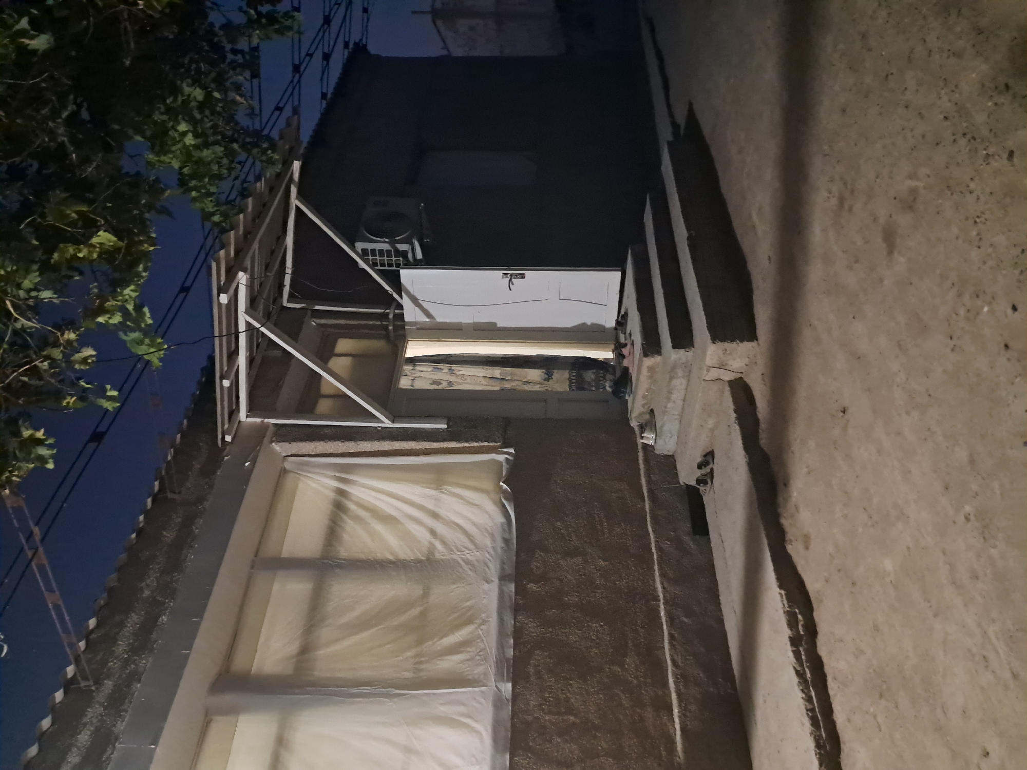 The image depicts the exterior of a small, single-story building at night, in Gurugli, Uzbekistan. The building has a light-colored, textured wall and a sloped roof covered with dark, corrugated material. A white door is ajar, revealing a patterned curtain inside. An air conditioning unit is mounted on the right side of the building. In front of the door, there are several concrete steps leading up to a paved area. A pair of dark-colored shoes and a white pair of shoes are placed on the steps, and a pair of slippers are near the doorway. A large window on the left side of the building is covered with white material, possibly a sheet or curtain, obscuring the interior. The ground in front of the building is dirt or gravel. Trees with green leaves are visible in the upper right portion of the image, and power lines crisscross the dark sky. The overall scene suggests a residential or utility structure in a somewhat rural or suburban setting.