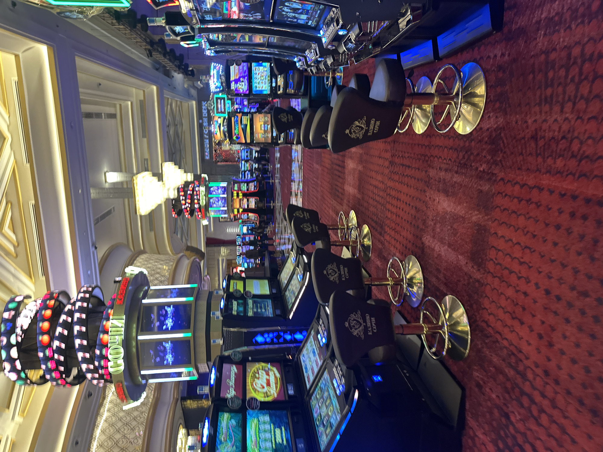 The image depicts the interior of a brightly lit casino, specifically located in Estosadok, Russia, as indicated by signage. The scene is dominated by rows of slot machines, their screens displaying various game graphics and neon lights. In the foreground, several black casino chairs with gold accents are neatly arranged in front of the machines, some with the logo "КАЗИНО СОЧИ" (Casino Sochi) visible on their backs. The floor is covered with a patterned red carpet. A large, multi-tiered cylindrical structure with flashing lights and a sign reading "СОЧИ" is visible in the background, further confirming the location and purpose of the establishment. The overall atmosphere suggests a lively and visually stimulating environment typical of a casino.