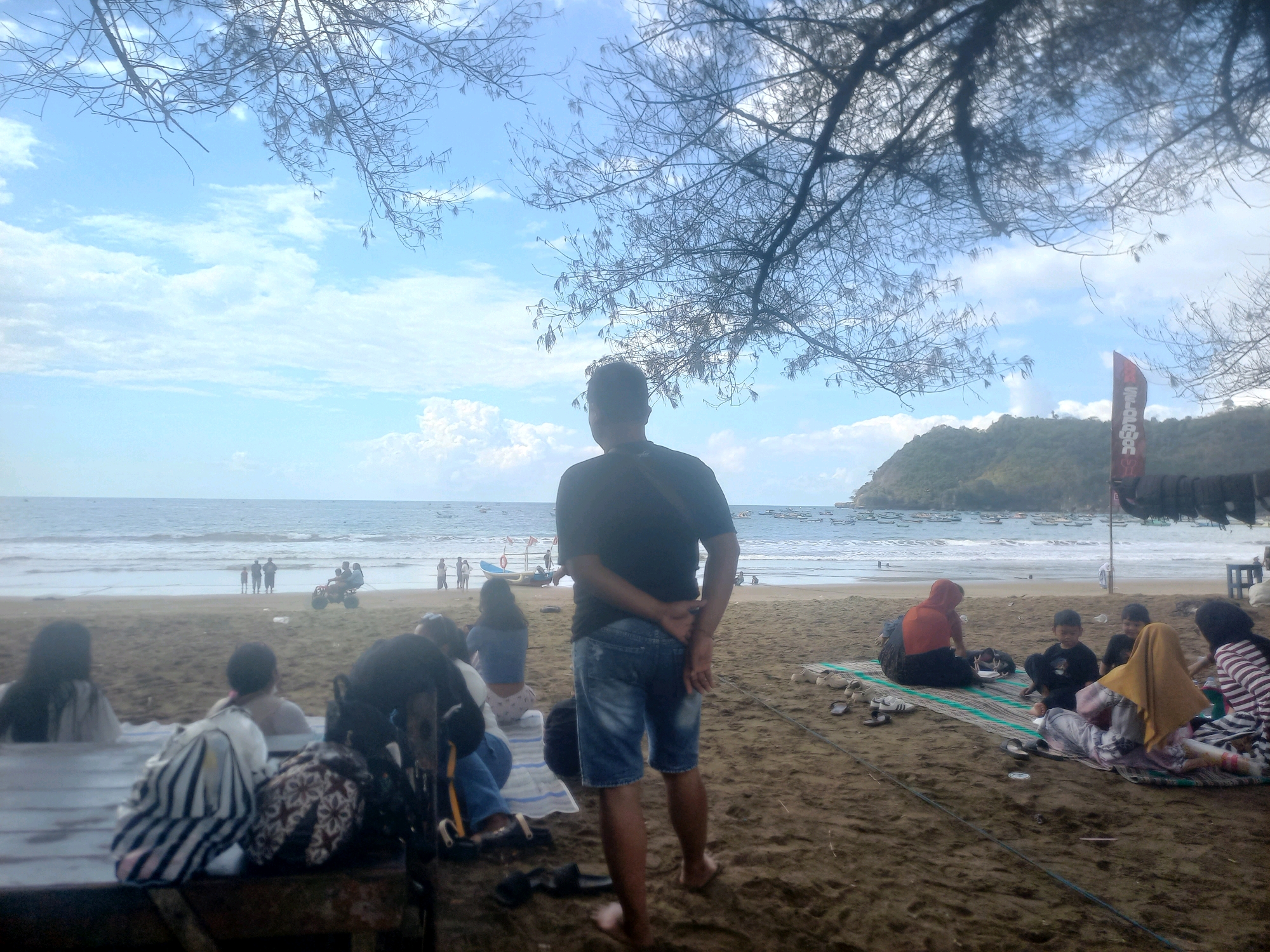 A group of people are enjoying a day at the beach. In the foreground, a man stands with his back to the camera, wearing a black t-shirt and denim shorts. To his left, a woman with long dark hair sits, wearing a light-colored top and dark pants. To the right, several other individuals are seated on mats on the sand. These include a person wearing an orange headscarf and a dark outfit, two young children, and a woman wearing a yellow headscarf and a striped shirt. The beach is sandy, with some scattered items like bags and footwear. In the mid-ground, the ocean stretches out to the horizon, with waves breaking on the shore. Several small boats are visible on the water. Further back, a sandy slope leads up to a vegetated hill. A red and white flag is planted on the beach. The sky above is blue with scattered white clouds. A branch of a tree hangs down from the top of the frame, partially obscuring the view. The scene appears to be a typical day at a coastal area in Indonesia.