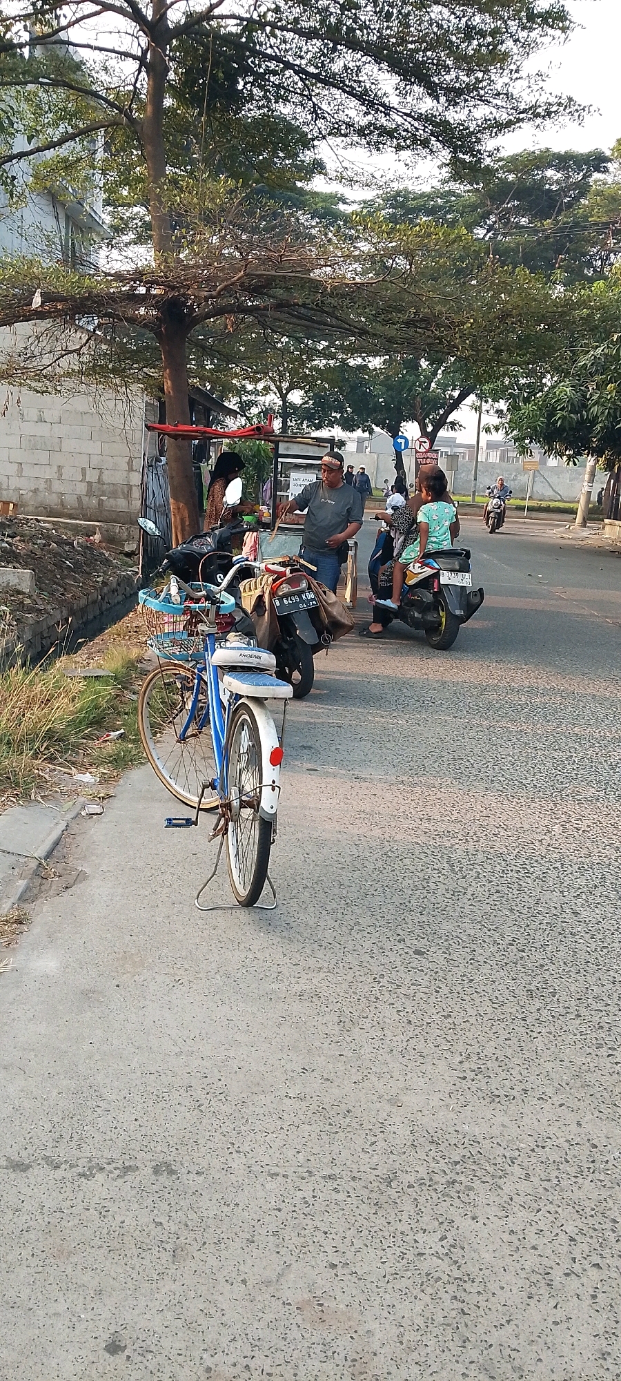 In Satriamekar, Indonesia, a street scene unfolds with a blue bicycle parked on the side of a road, its front basket filled with various items. Behind the bicycle, a man in a grey t-shirt and blue jeans stands near a food cart, holding a long skewer. A woman wearing a headscarf is visible at the food cart. Further down the road, two motorbikes are present. On one, a woman in a white and green patterned dress sits behind a person wearing a black head covering, who appears to be riding the motorbike. Another motorbike is parked nearby. In the background, a few people are walking, and trees line the street. The environment suggests a warm, sunny day.
