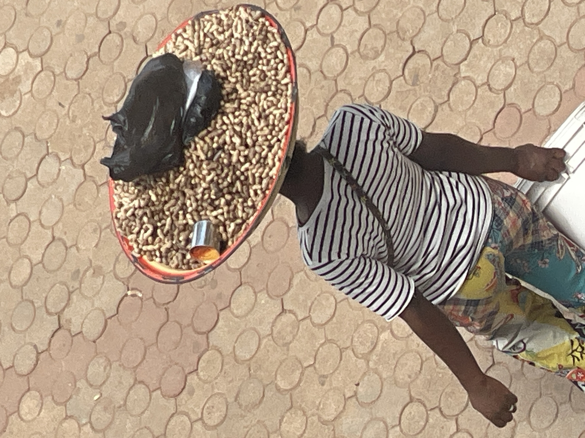 A person balances a large, shallow bowl overflowing with peanuts on their head. The bowl is red with a dark rim. A black plastic bag is nestled amongst the peanuts in the bowl, and a small metal container, possibly a cup or scoop, is visible near the edge. The person is wearing a horizontally striped t-shirt with black and white stripes, and colorful patterned pants. A strap from a bag or accessory crosses their chest. They are walking on a paved surface made of reddish-brown interlocking stones. The scene appears to be an outdoor market or street in Jos, Nigeria.
