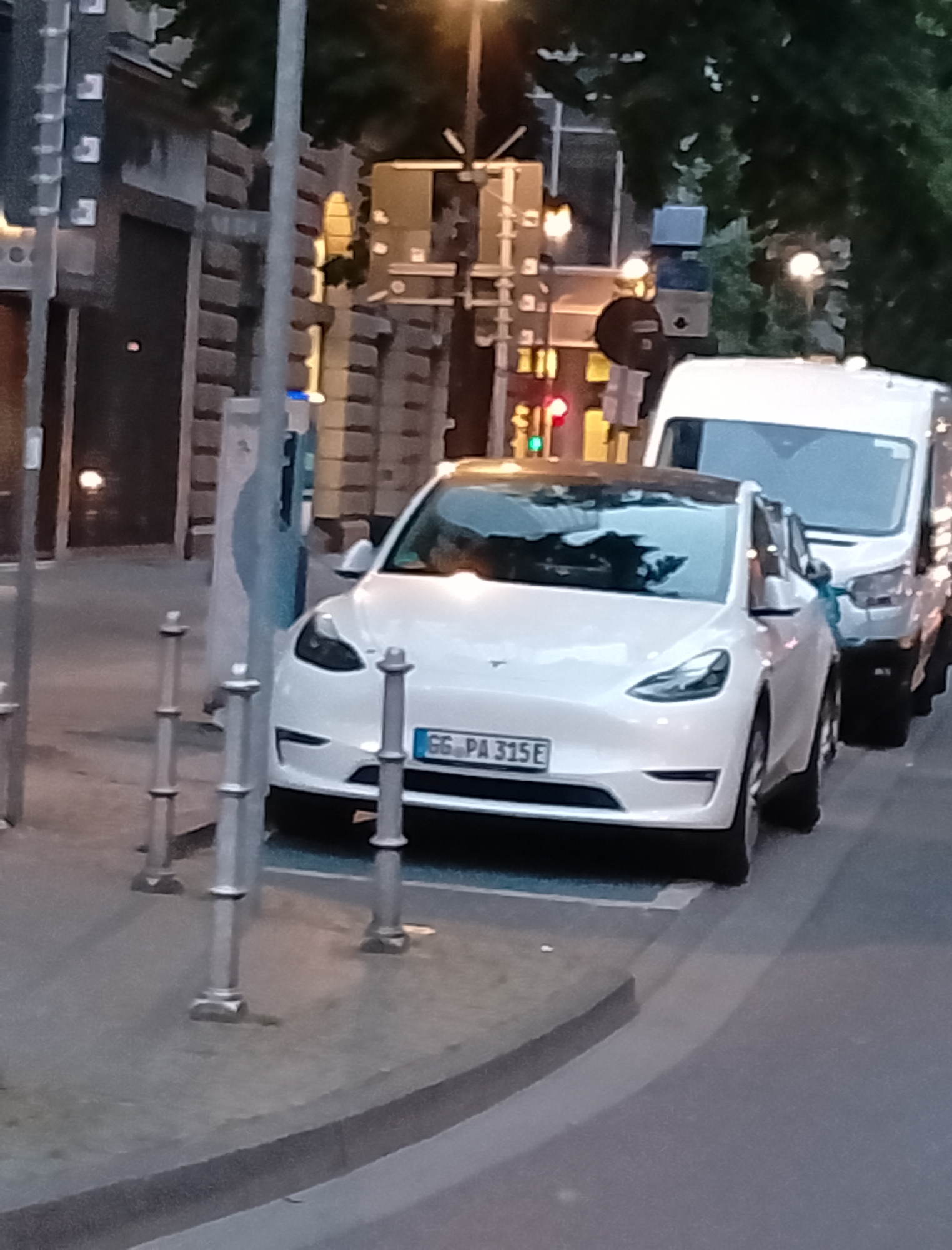 A white Tesla Model Y is parked on a city street. The car has a license plate that reads "GG PA 315E". Behind the Tesla, a white van is also parked. The street appears to be in a European city, as indicated by the license plate format and the style of the buildings in the background. The scene is illuminated by streetlights and traffic signals. There are trees and pedestrian bollards present.