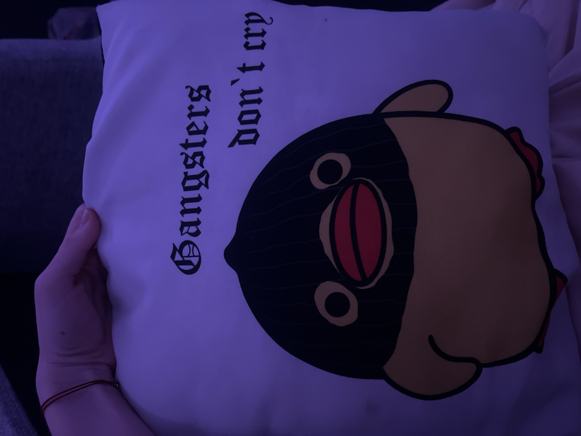 A person is holding a square white pillow with a cartoon penguin printed on it. The penguin is predominantly black, with yellow accents around its head. It has large, cartoonish eyes and a red mouth. To the left of the penguin are the words "Gangsters don't cry" in a Gothic font. The person's hand and wrist are visible, with a red string bracelet around the wrist. The background appears to be a grey couch. The location is not discernible.