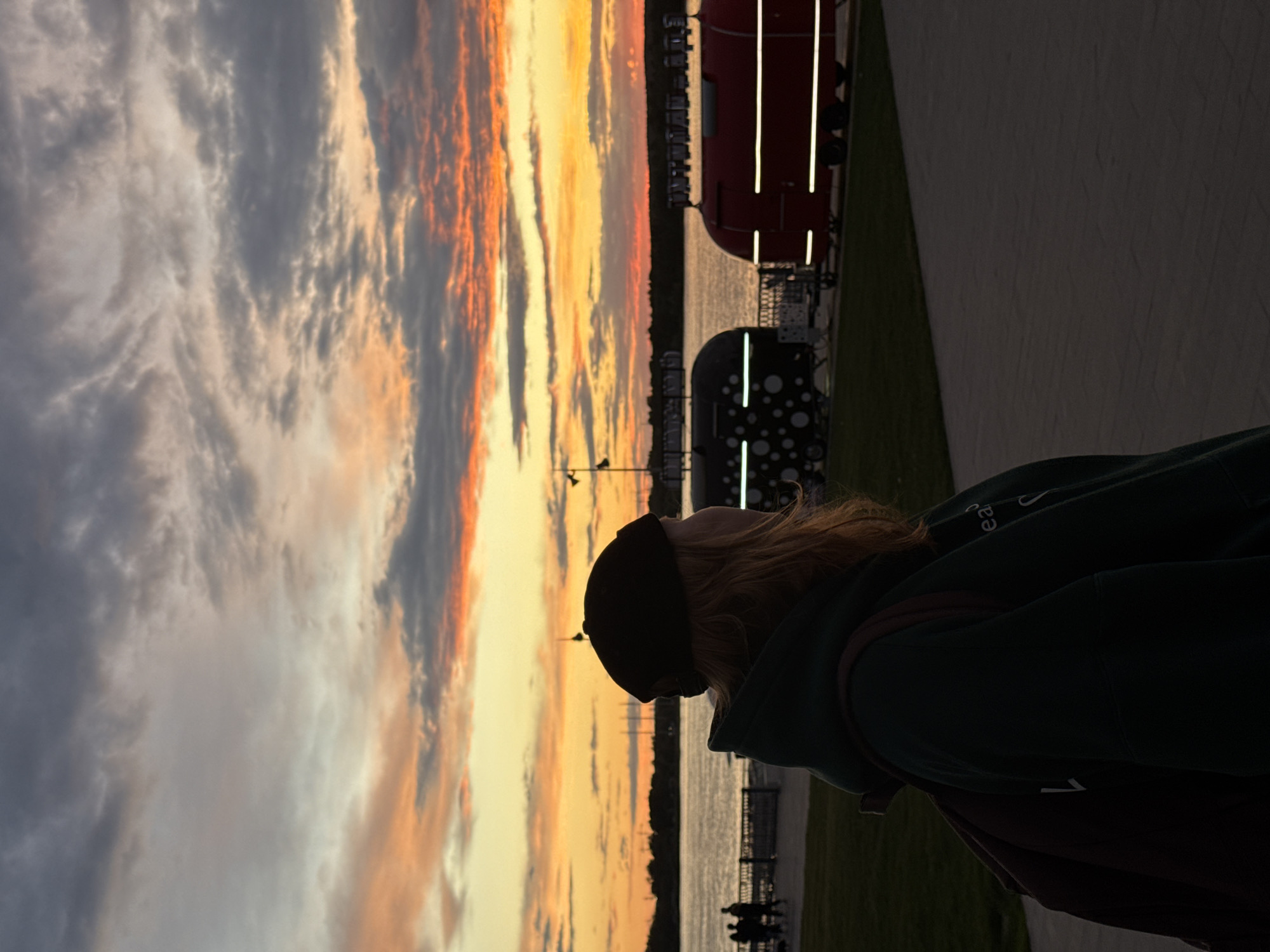 The image shows a woman with blonde hair and a black cap looking towards a sunset over the Kama River in Perm, Russia. The sunset displays a mix of orange, yellow, and gray clouds. The woman is wearing a dark green hoodie and carrying a brown backpack. In the background, along the riverfront, there are illuminated art objects, including a red structure with vertical white stripes and a black one with white polka dots. The scene also includes a walkway and a grassy area near the riverbank.