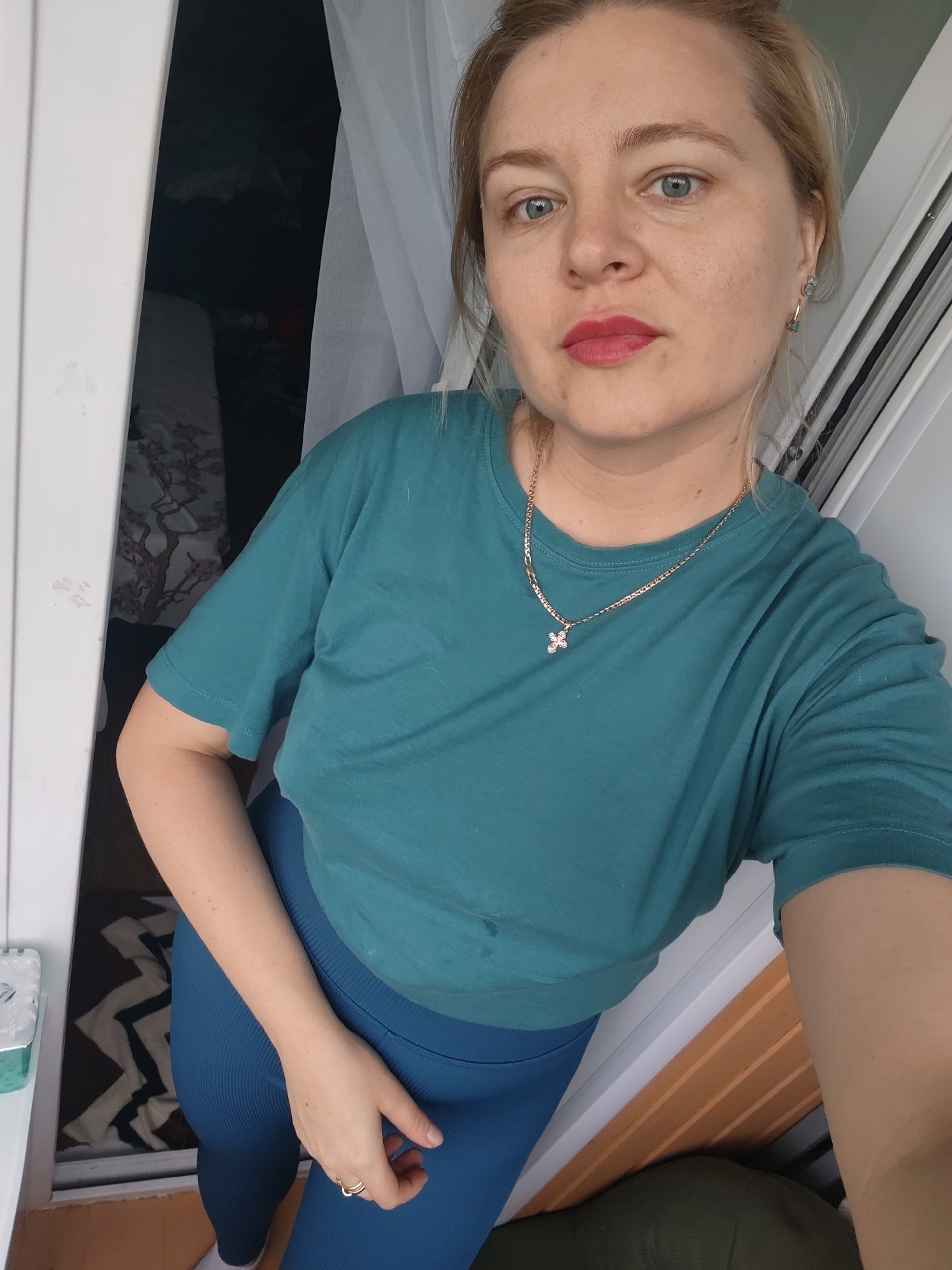 A woman with blonde hair and blue eyes is captured in a selfie, standing indoors near a doorway. She is wearing a teal t-shirt and blue leggings, accessorized with a gold necklace featuring a small cross pendant and a ring on her right hand. The background reveals a glimpse into a room with clothing or fabric visible, as well as a window with white curtains. The overall lighting suggests an indoor setting. The provided information places this scene in Izhevsk, Russia.