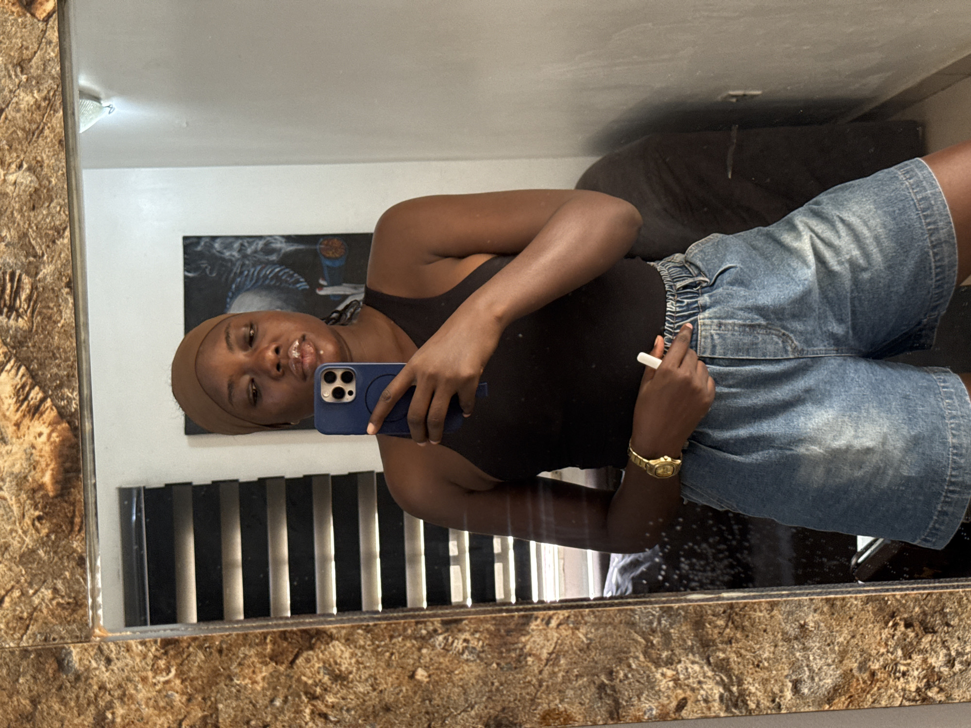A young woman is depicted in a mirror selfie, taken indoors, likely within a home in Abuja, Nigeria. She stands upright, facing slightly towards her right, capturing her reflection in a rectangular mirror with a textured, possibly stone-like frame. The woman has dark skin and is wearing a light brown head wrap or cap, a fitted black sleeveless top, and light blue denim shorts with an elasticated waistband.
She holds a blue smartphone with a case in her left hand, angling it to take the photograph, with her eyes looking directly towards the camera in the reflection. A gold-colored watch adorns her right wrist, and her right hand holds a slender white object, possibly a pen or vape, resting near the waist of her shorts. Her expression is neutral to gently smiling.
The background reflected in the mirror includes a plain white wall. On the wall, a dark-toned artwork or painting featuring abstract elements, including what appears to be a face with dreadlocks or waves and splashes of blue and orange, is visible. Below this, black horizontal blinds or slatted decor can be seen. A dark-colored sofa or bed is partially reflected on the right side of the frame. A light fixture is also visible on the ceiling in the upper left corner of the reflected room. The overall scene suggests a casual and personal space during daytime.