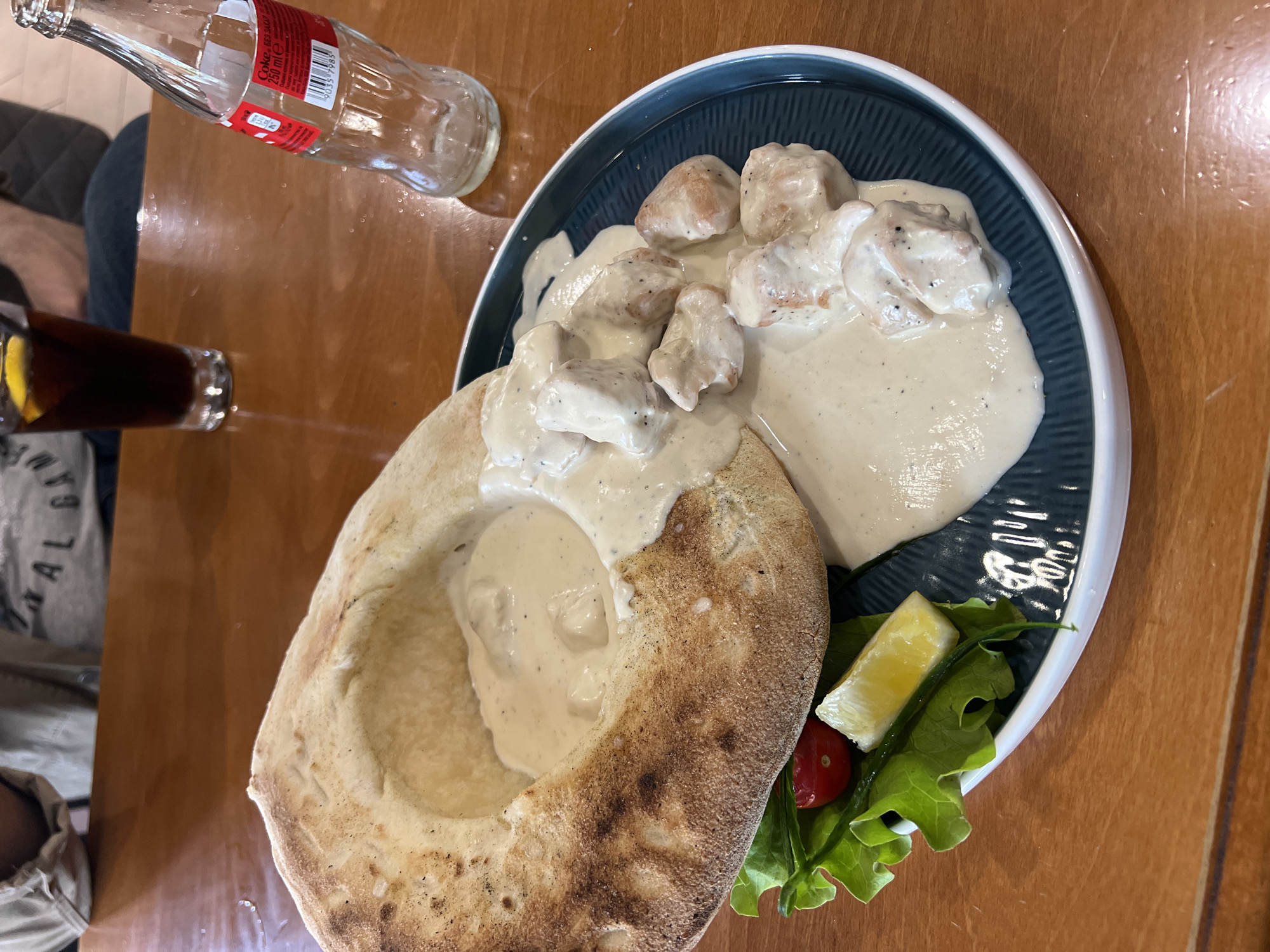 A table scene shows a meal in Gabrovo, Bulgaria. A round bread bowl filled with a creamy sauce sits on a dark blue-green plate with white trim. The plate also holds pieces of chicken in the same sauce, a slice of lemon, lettuce, and a cherry tomato. A bottle of Coca-Cola and a glass of cola are on the wooden table. The scene suggests a restaurant setting.